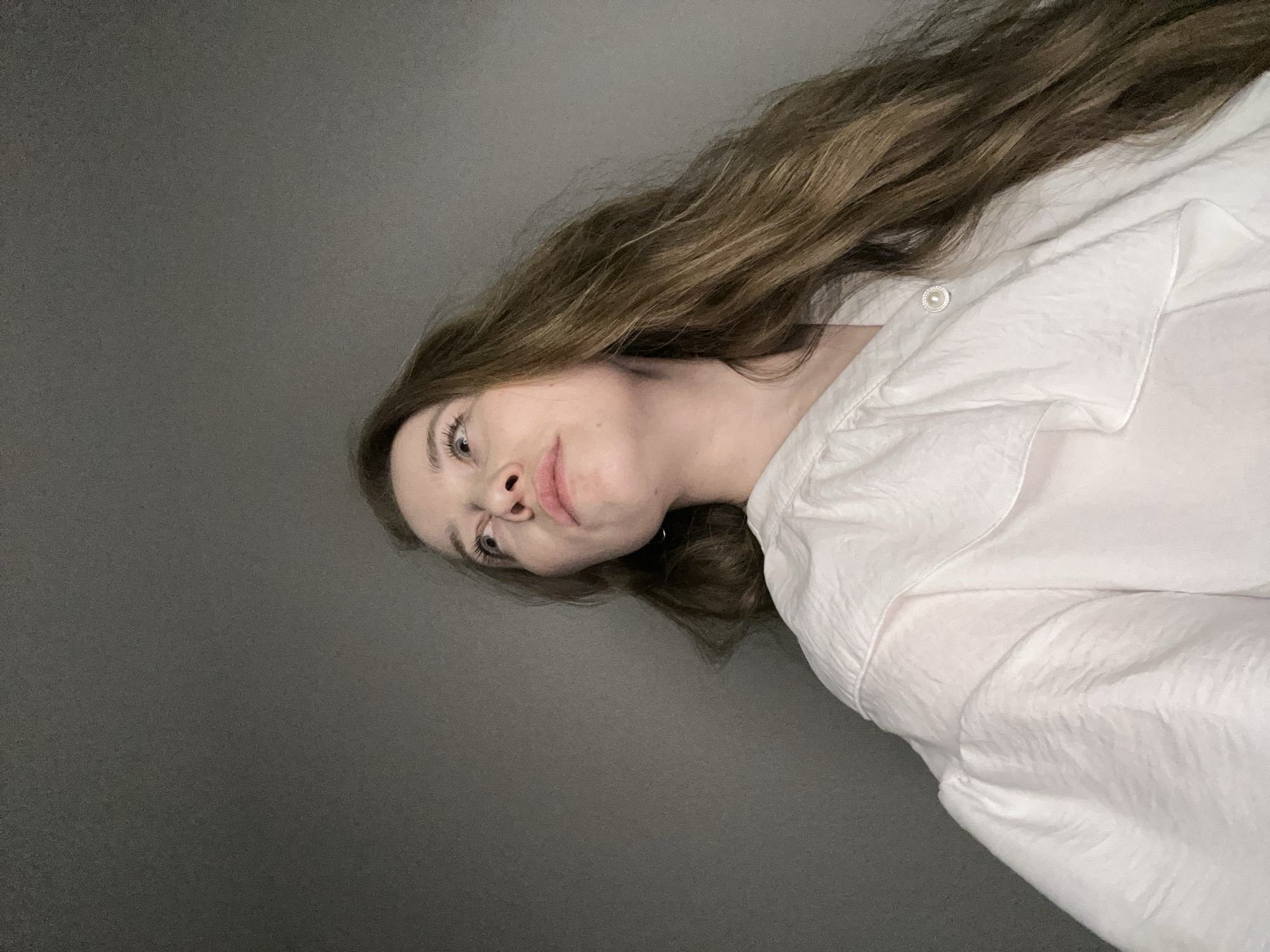 A woman with long brown hair is seen in a close-up, angled shot. She is wearing a white collared shirt, which appears to be made of a satin-like material and has a visible button. The background is a solid, dark gray. The image does not show any features indicative of the city of Saint Petersburg, Russia. Therefore, location information cannot be inferred from the visual data.