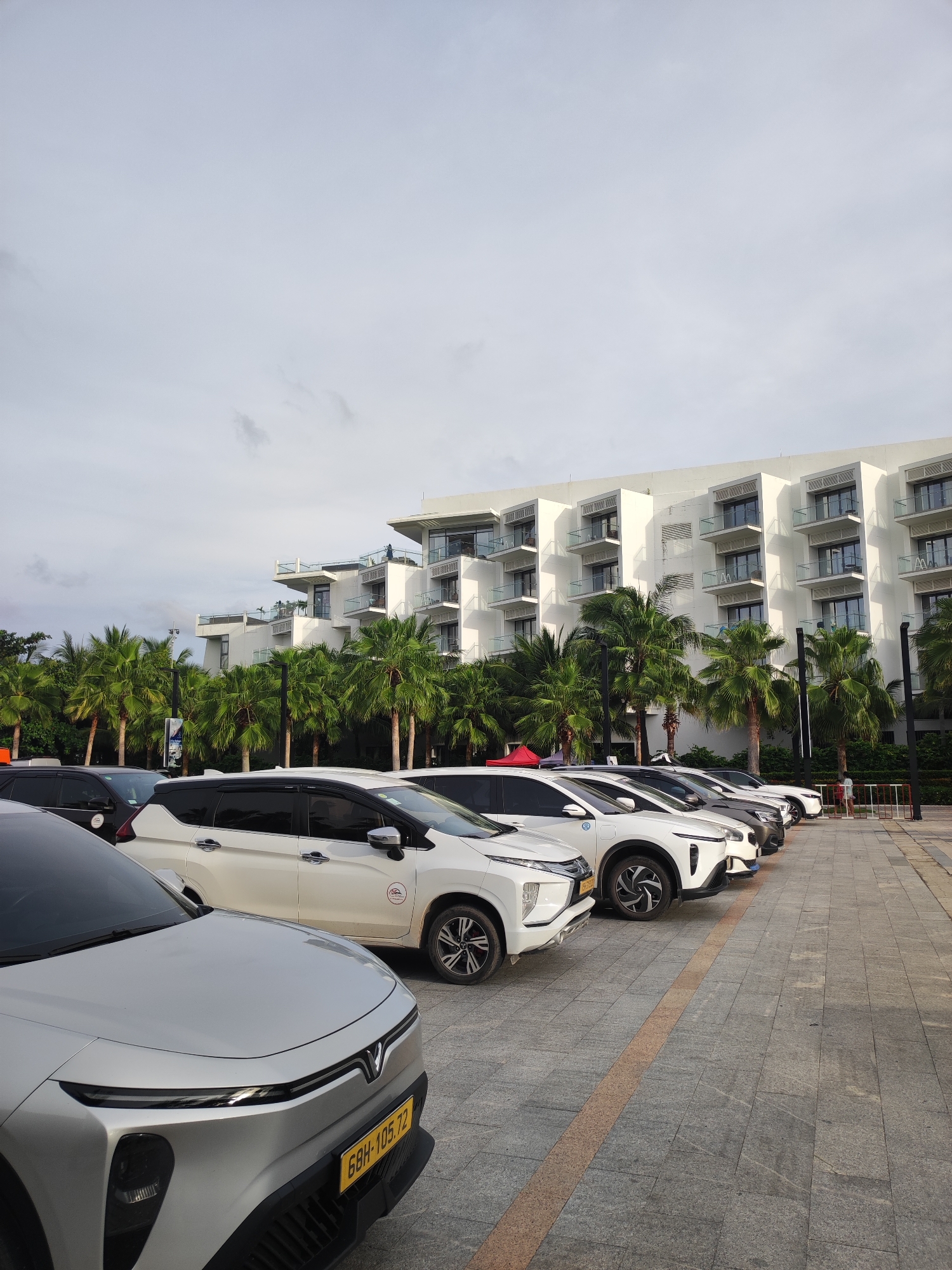 This is a wide-angle, eye-level shot of a row of parked cars in front of a multi-story building with a modern, white facade and numerous balconies, situated in Phu Quoc, Vietnam. The building is surrounded by tall, lush palm trees. The sky is overcast and pale. The cars are all facing to the right and are arranged in a parking lot paved with grey and tan tiles. In the foreground, the front of a silver electric vehicle with a yellow license plate bearing the characters "68H-105.72" is visible. Behind it, other vehicles, including a white car with a black roof and a smaller white SUV with a red circular decal on its side, are parked in a line. A few people are visible in the background near the building, including one person walking on the sidewalk. The overall scene suggests a resort or hotel environment due to the architecture, palm trees, and the presence of multiple cars.