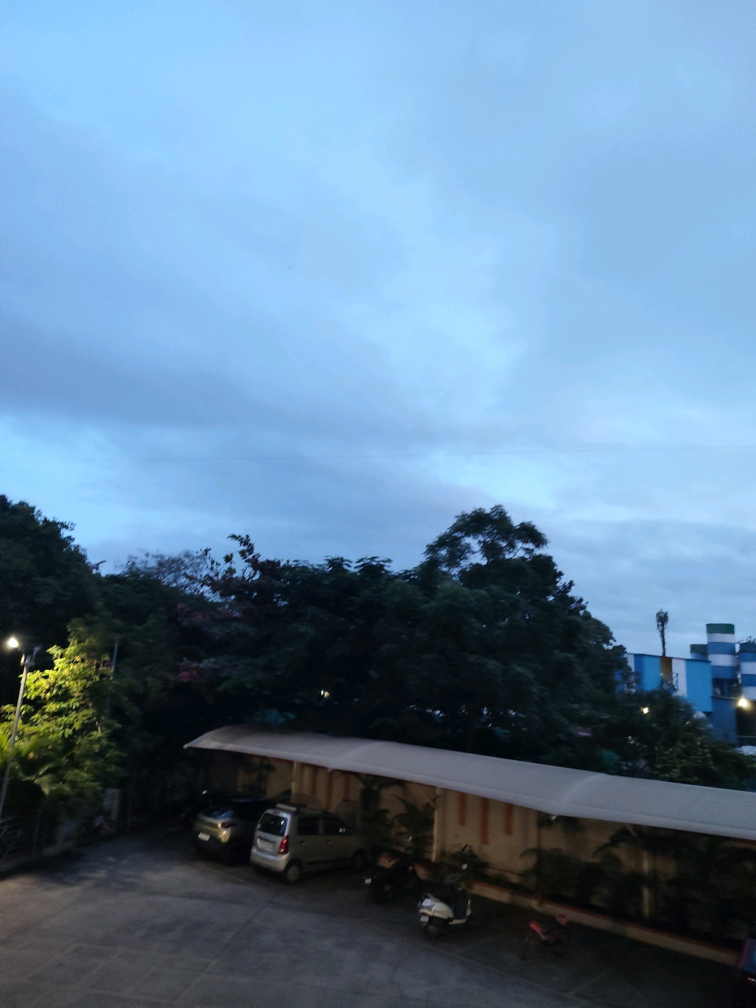 The image depicts an outdoor scene at dusk or dawn, with a cloudy sky dominating the upper portion of the frame. The lower section of the image shows a paved area with several parked vehicles, including a silver hatchback and a dark-colored sedan. Adjacent to the parking area is a building with a light-colored, curved awning supported by pillars. Behind the building, a line of trees and foliage is visible, with some trees appearing dense and dark, while others are lighter and illuminated by a street lamp on the left. In the background, a tall telecommunications tower and parts of a multi-story building with blue and white sections are discernible. The overall ambiance suggests an urban or suburban setting in Anandnagar, India, likely a commercial or residential complex. There are no people visible in the image, and the focus is on the environment and stationary objects.