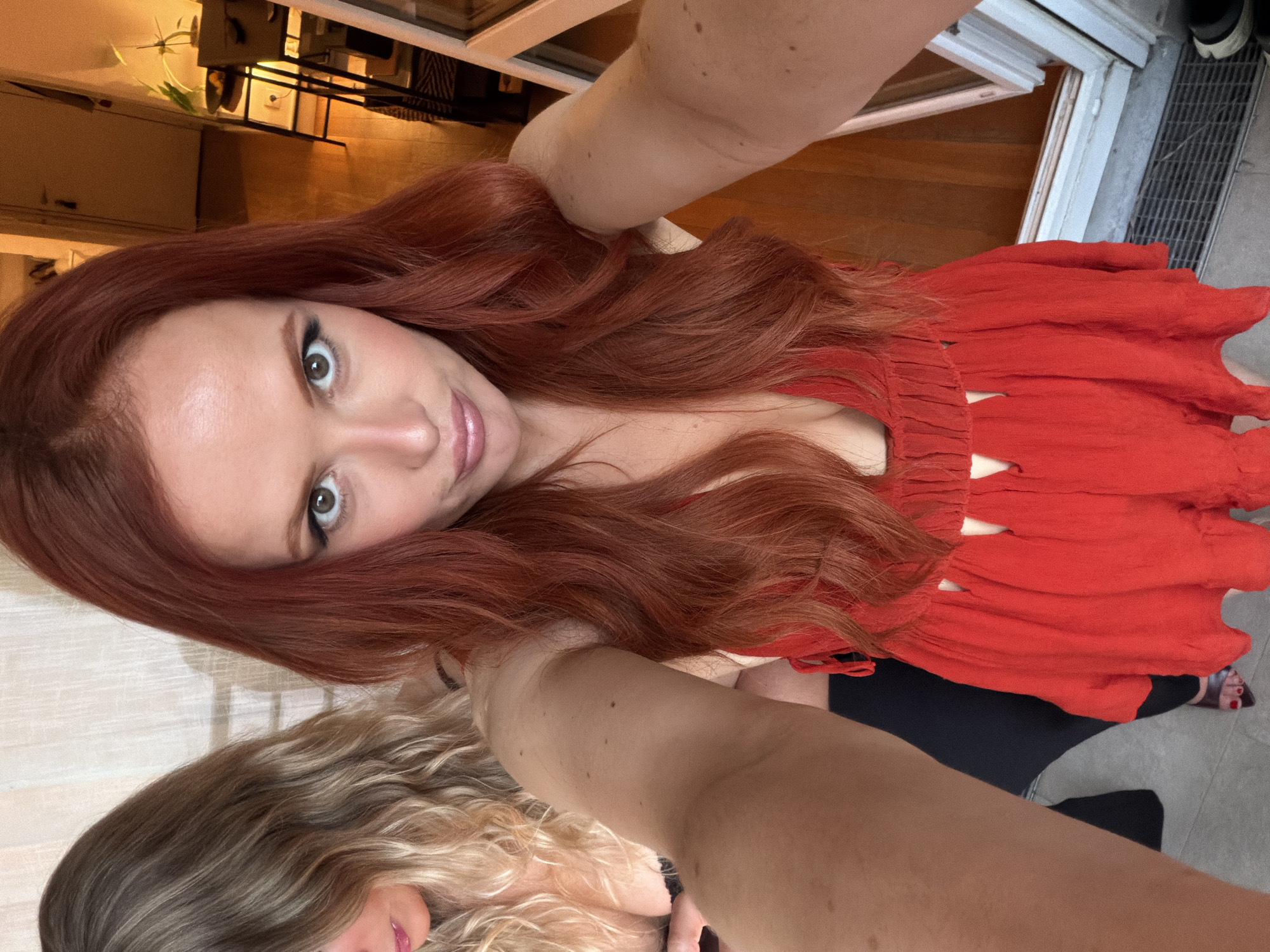 A selfie taken from above, angled, features two women indoors. The woman in focus has long, wavy, auburn hair and green eyes; she wears a red dress with cutout details. The second woman, partially visible in the lower left corner, has blonde, wavy hair. The background is a room with wooden floors. A doorway leads to what appears to be an exterior view of a sidewalk or street with a grate and a glimpse of footwear. Details such as furniture and lighting fixtures are also visible. The location is potentially Paris, France, though without more information, one cannot be definitive.