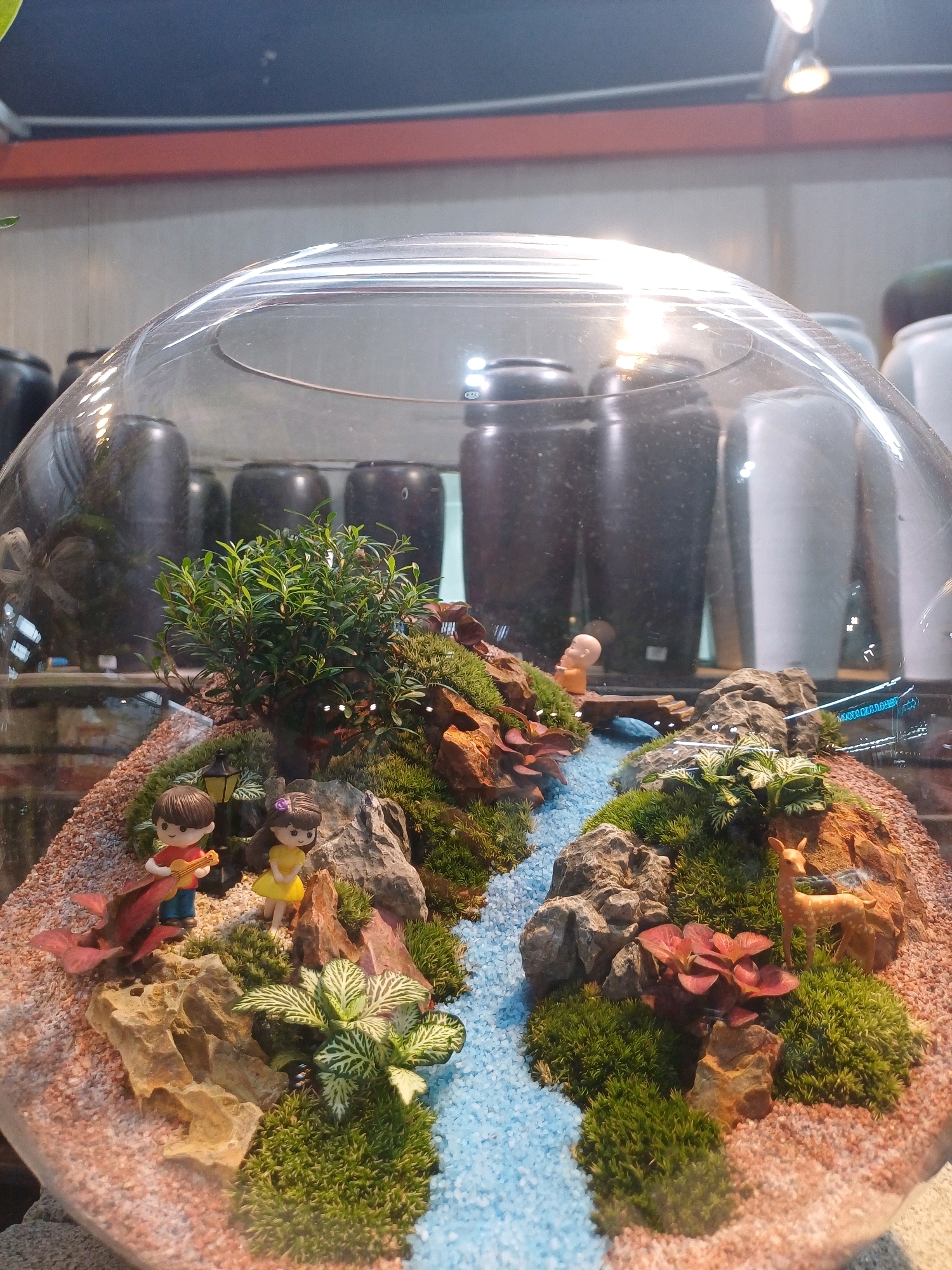 The image shows a miniature diorama inside a glass dome, placed in what appears to be a retail or display setting. The diorama depicts a small landscape with a flowing blue stream, mossy hills, rocks, and various miniature plants. Two figurines of children, one playing a guitar and the other standing, are positioned on the left side of the diorama. A tiny deer figurine is seen on the right. In the background, large dark cylindrical objects are visible, possibly vases or storage containers, along with some white objects. A faint reflection of lights and some text on the glass dome are also present. The overall impression is of a decorative item displayed in a well-lit indoor environment. There is no specific indication of its location within Kuala Lumpur, Malaysia.