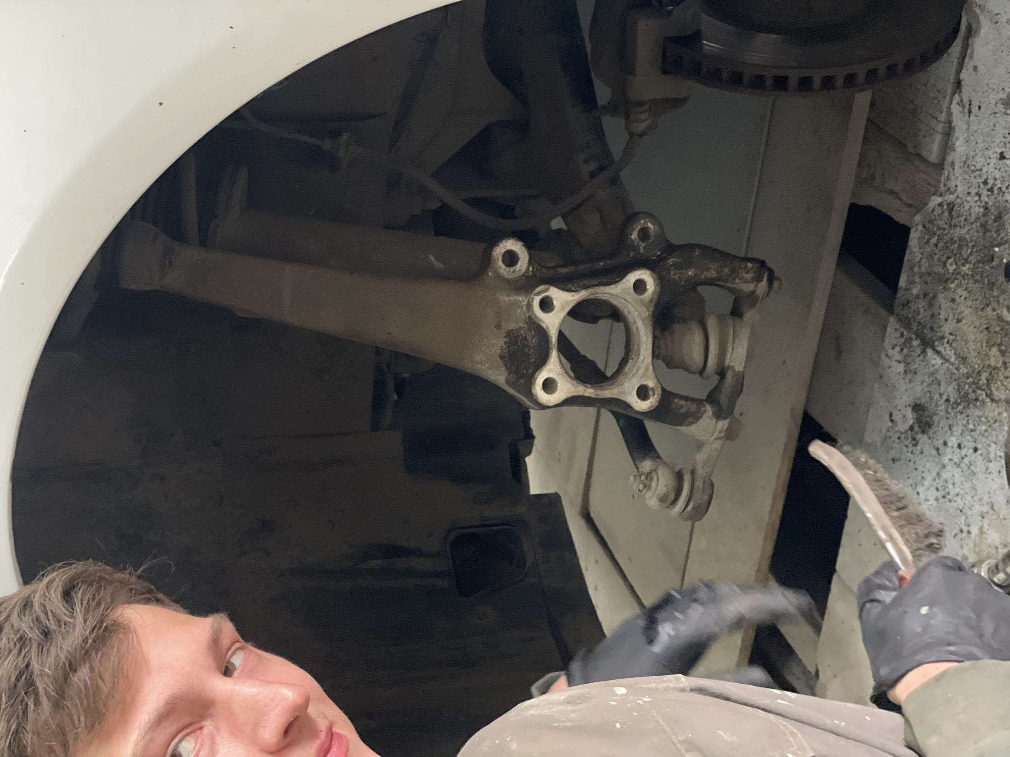 A man with short brown hair is partially visible in the lower left corner of the image. He appears to be lying on his back, looking upward towards a vehicle's wheel well. The wheel well contains a vehicle component, likely a suspension or steering part, that is grimy with dirt and grease. A gloved hand holding a brush is cleaning the component. The setting seems to be a repair shop or garage. There is no way to prove the location in the city of Минино, Russia.
