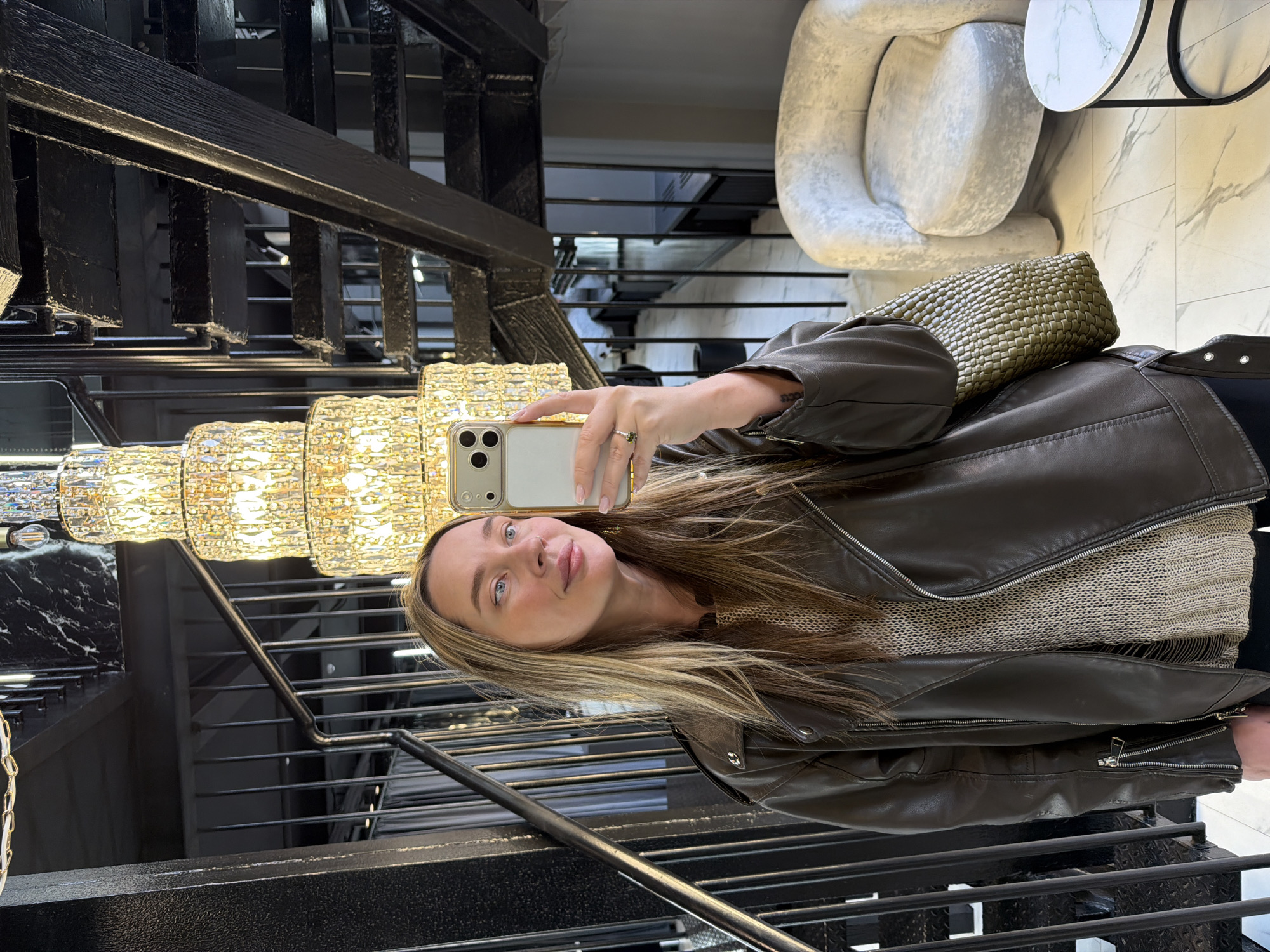 A young woman is captured taking a selfie with her smartphone in a luxurious indoor setting. She has long, straight blonde and light brown hair and light blue eyes, with a slightly pouting or smirking expression. She is wearing a dark brown or olive green leather-like moto jacket over a light beige or off-white knitted sweater. On her right hand, holding the white-backed smartphone with a gold-trimmed clear case (featuring a triple camera array), she wears a ring with a prominent green stone.
The background is rich with detail, indicating an opulent environment. To her left, a large, multi-tiered chandelier with numerous shimmering, possibly crystal or glass, elements provides warm illumination. Below and behind the chandelier, a dark, industrial-style staircase with black metal railings descends. Behind the woman's right shoulder, an olive green or khaki-colored handbag with a distinct woven texture rests. Further in the background, a large, light-colored, abstractly shaped lounge chair or sculpture is visible, alongside a small, round table with a white and grey marble-patterned top. The floor or wall behind these elements also features a white marble pattern. The overall ambiance suggests a high-end establishment such as a modern hotel lobby or an upscale private residence, located in Moscow, Russia. The setting is well-lit, likely during the daytime or in a brightly illuminated interior. No discernible text is visible in the image.