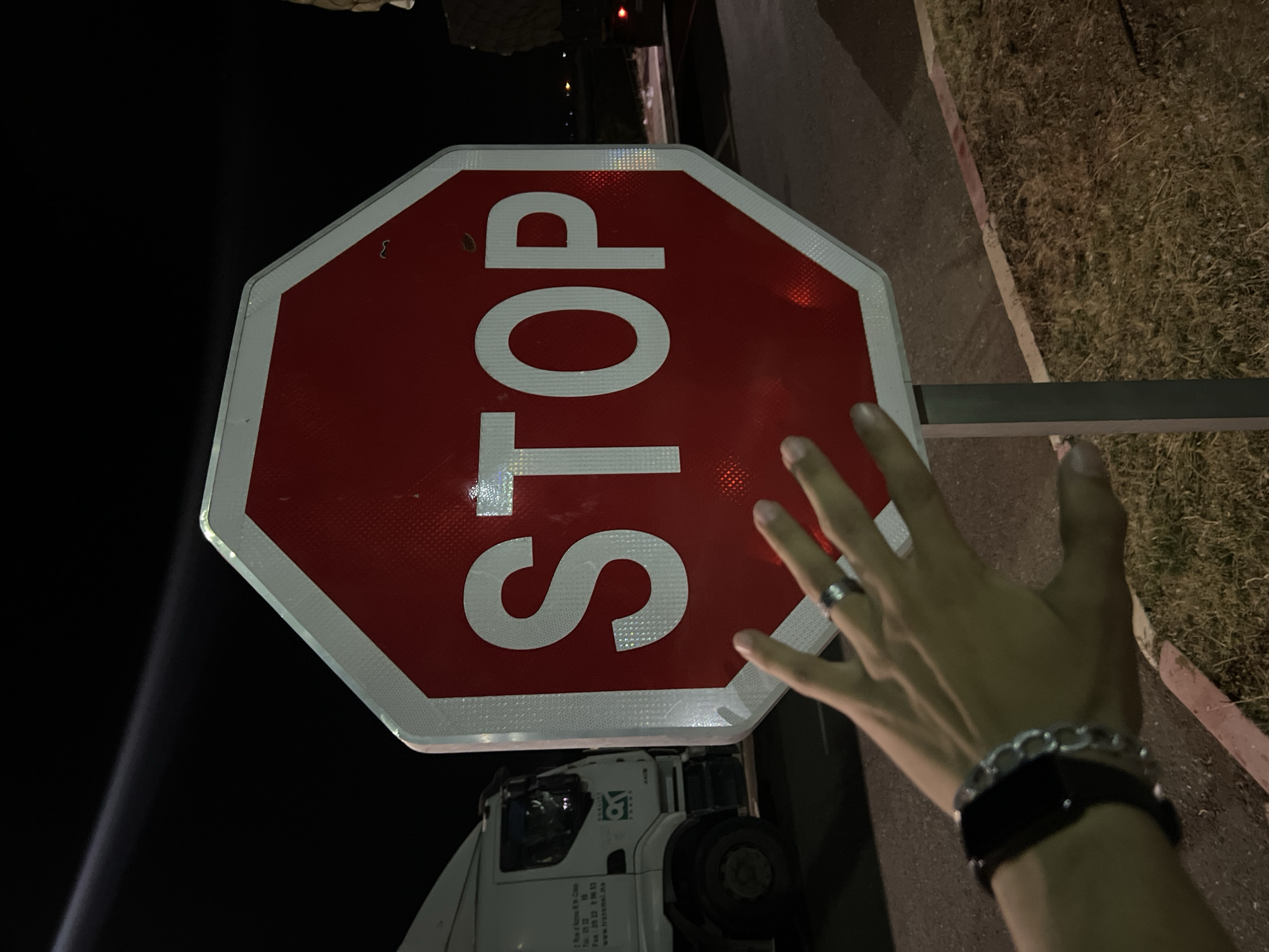 A person extends their hand towards a red and white STOP sign in Jbilate, Morocco. The sign is mounted on a pole. A truck is visible below the sign. The surrounding environment includes pavement, grass, and darkness. The person is wearing a ring, a chain bracelet, and a watch.