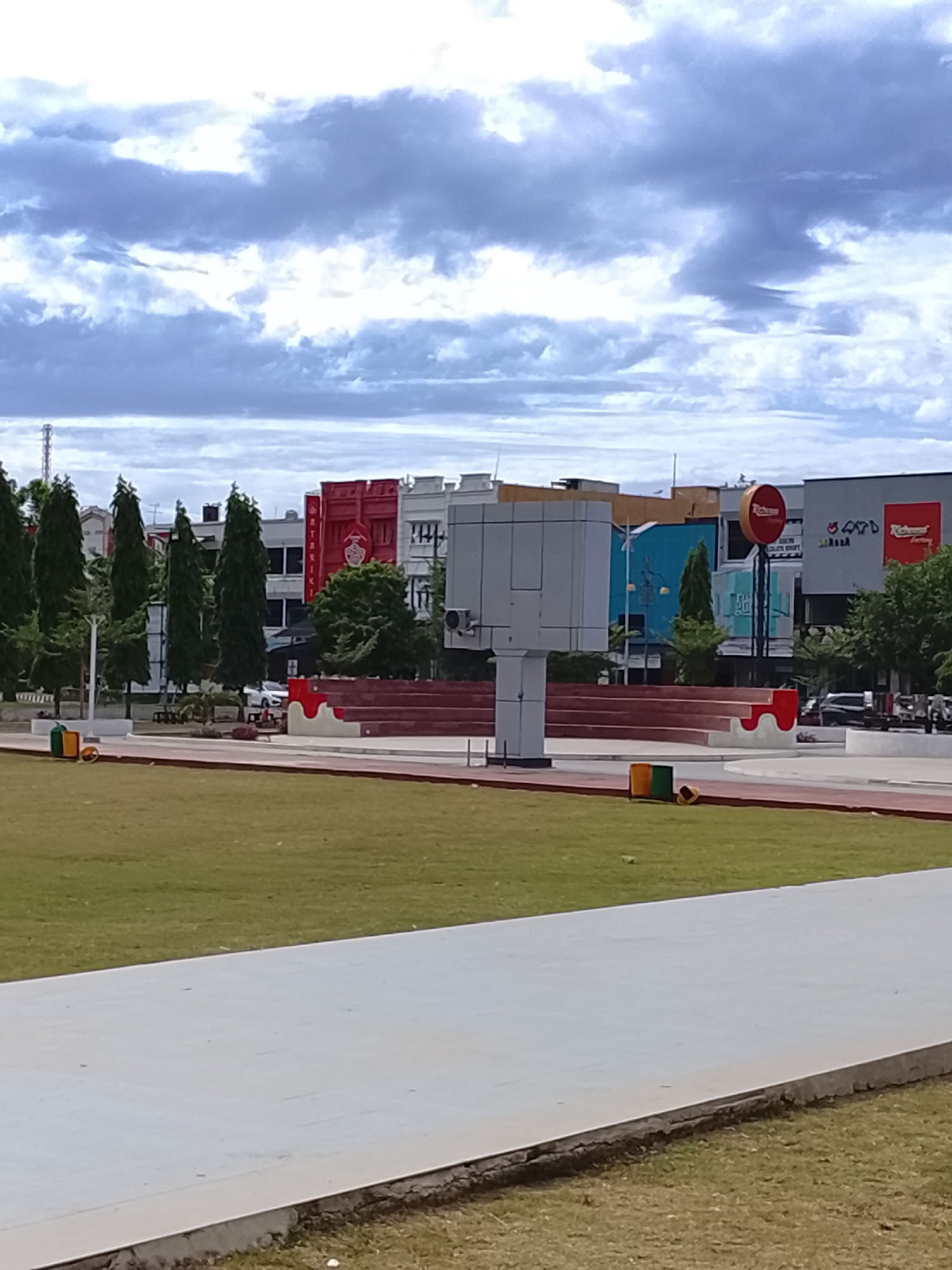 An eye-level shot captures a spacious urban public park or plaza under an overcast sky in Lhokseumawe, Indonesia. In the foreground, a light gray concrete pathway runs horizontally, bordering a well-maintained green grassy area. A narrow red-orange track or border is visible beyond the pathway, suggesting an athletic or recreational space.
The midground features a red tiered seating structure, resembling an amphitheater, with white accents. To the left of this structure, several tall, slender evergreen trees line a paved area, behind which cars are partially visible. A prominent white, multi-segment structure stands near the center, possibly a monument or a light fixture. Yellow, green, and orange trash cans are scattered near the pathway.
In the background, a row of multi-story buildings forms the cityscape. One building on the left features a striking red facade with white text, where "BINTANG" and "RIKI" are discernible, along with a crest-like logo. Further to the right, a large red circular sign bearing the partial word "RICH" (likely "Richest" or similar) stands tall, part of what appears to be a fast-food restaurant. Adjacent buildings display various colors, including blue and gray, with some indistinct signage. The sky is largely covered by thick, light gray clouds, indicating an overcast day. No people are visible, giving the scene a quiet and expansive feel.