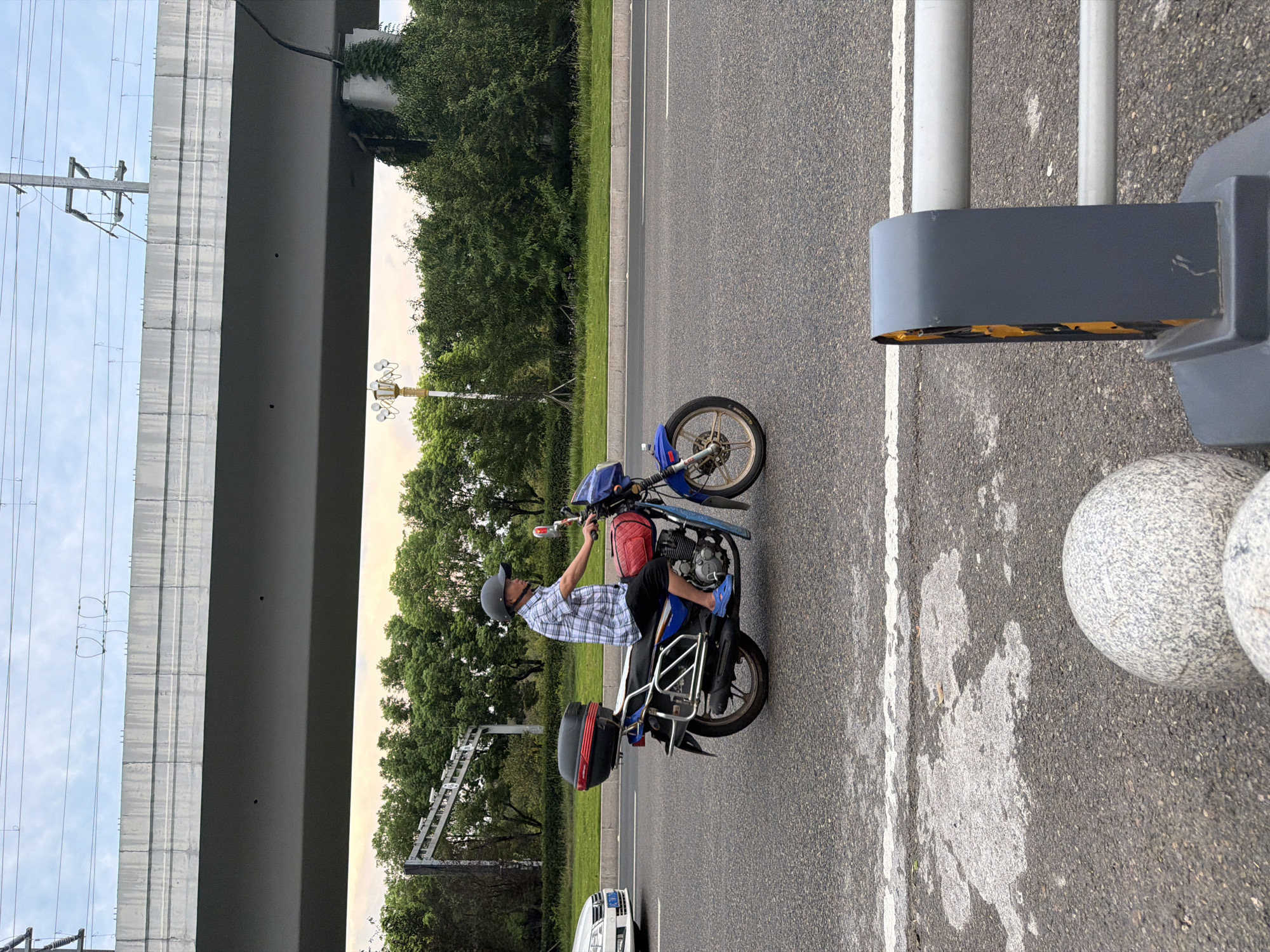 In Jiangjin, China, a man wearing a plaid shirt and a dark helmet rides a blue and red motorcycle on a paved road. The motorcycle has a black storage box attached to the rear. The man is wearing blue sandals. To the left of the motorcycle, a white car is visible. In the background, there are trees and a large overpass. The sky is overcast. In the foreground, parts of a grey barrier and two round granite bollards are visible.