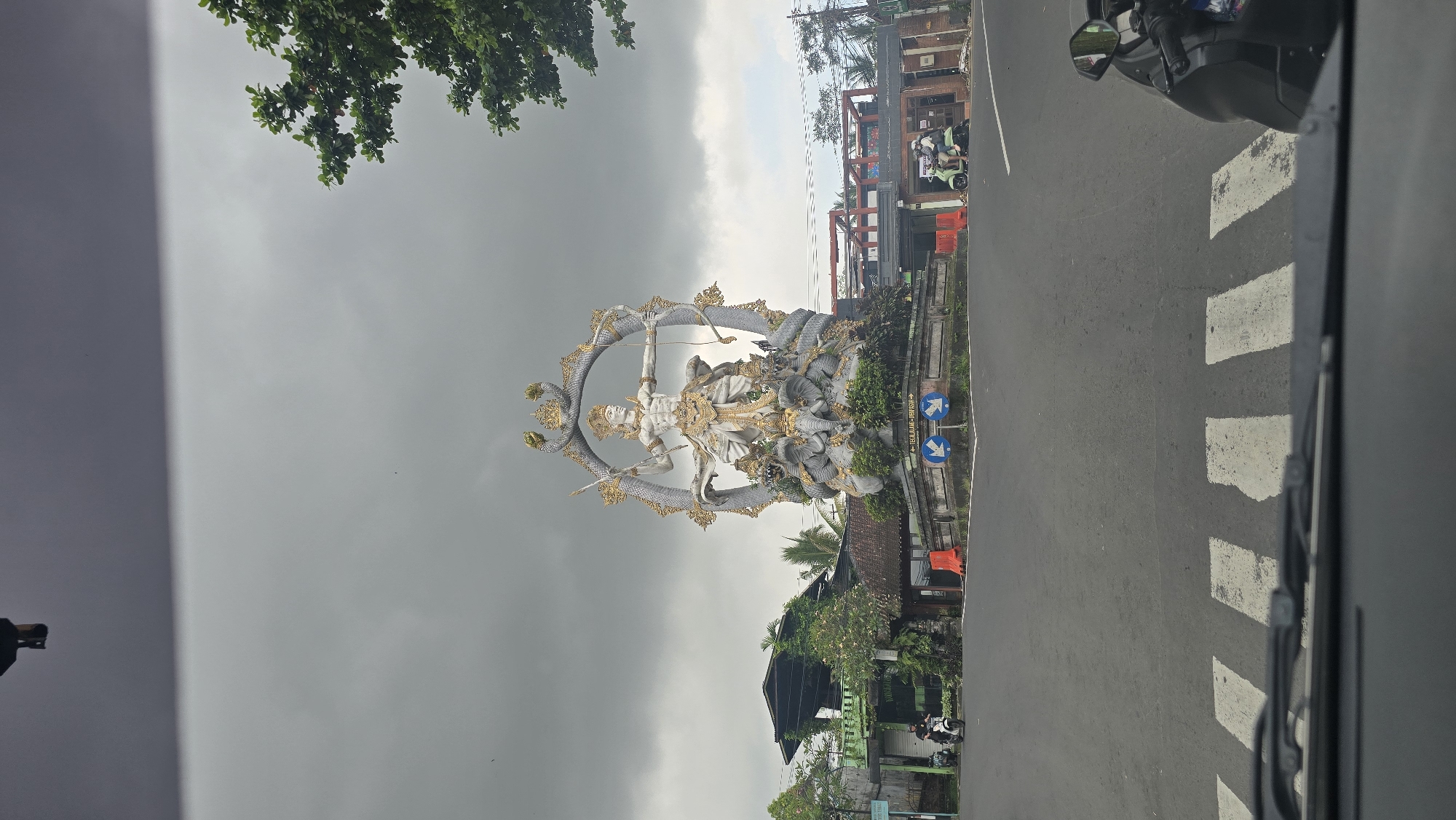 The image shows a statue of a Hindu deity holding a bow and arrow, encircled by a snake, located in a roundabout in Ubud District, Indonesia. The statue is made of white stone with gold accents and surrounded by lush greenery. In the background, there is a street with buildings and a cloudy sky. The photograph appears to be taken from inside a vehicle, possibly a car, with a portion of the vehicle's interior visible in the foreground. Pedestrian crosswalk markings are visible on the road.