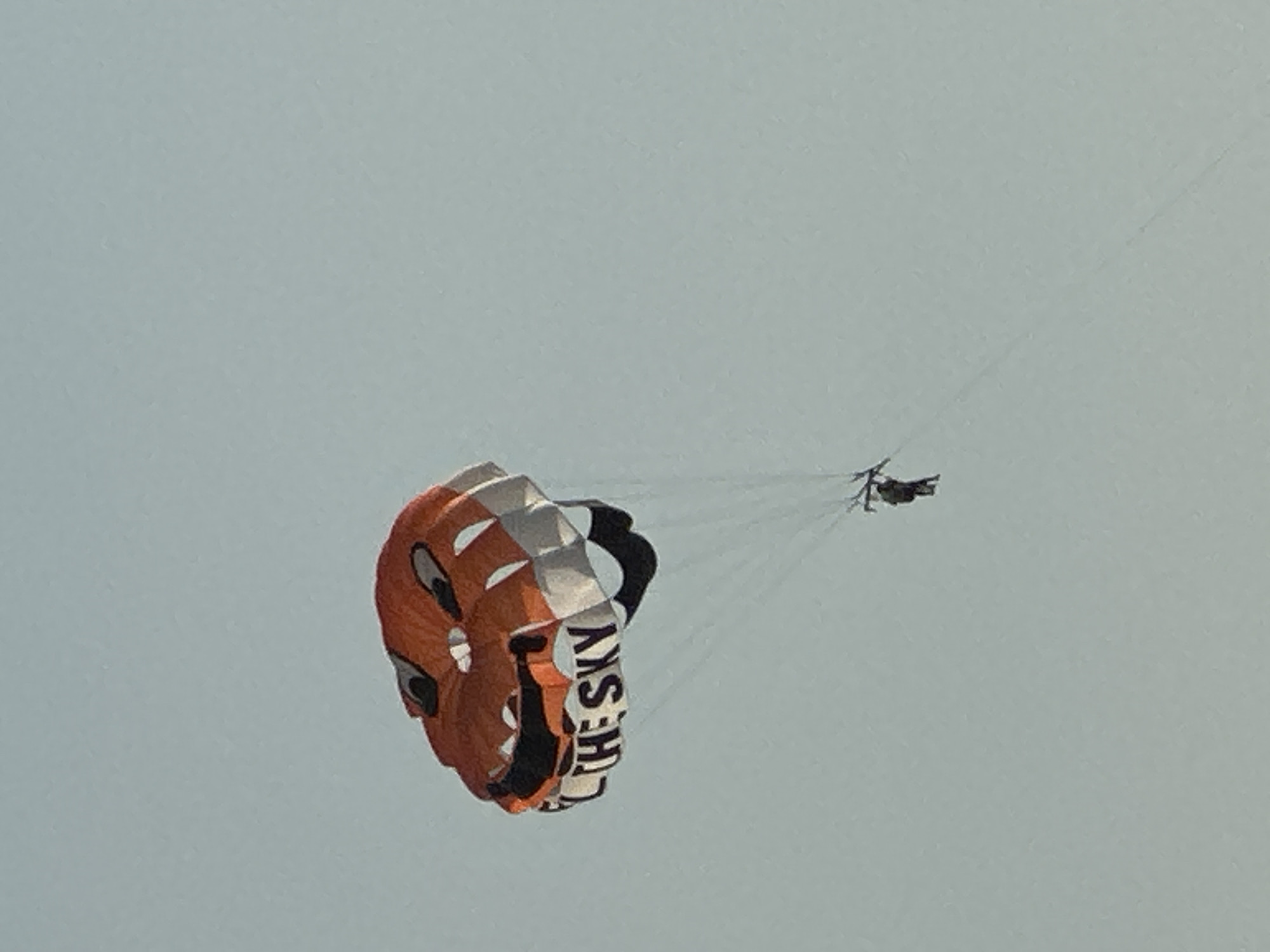 An orange and white parachute shaped like a jack-o'-lantern's face is being used for parasailing. The parachute has black eyes and a mouth, and the words "FLY THE SKY" are printed in black letters on the lower portion of the parachute. A person is suspended beneath the parachute, tethered by lines. The scene takes place in the sky above Boreti, Montenegro. The sky is clear and light blue.
