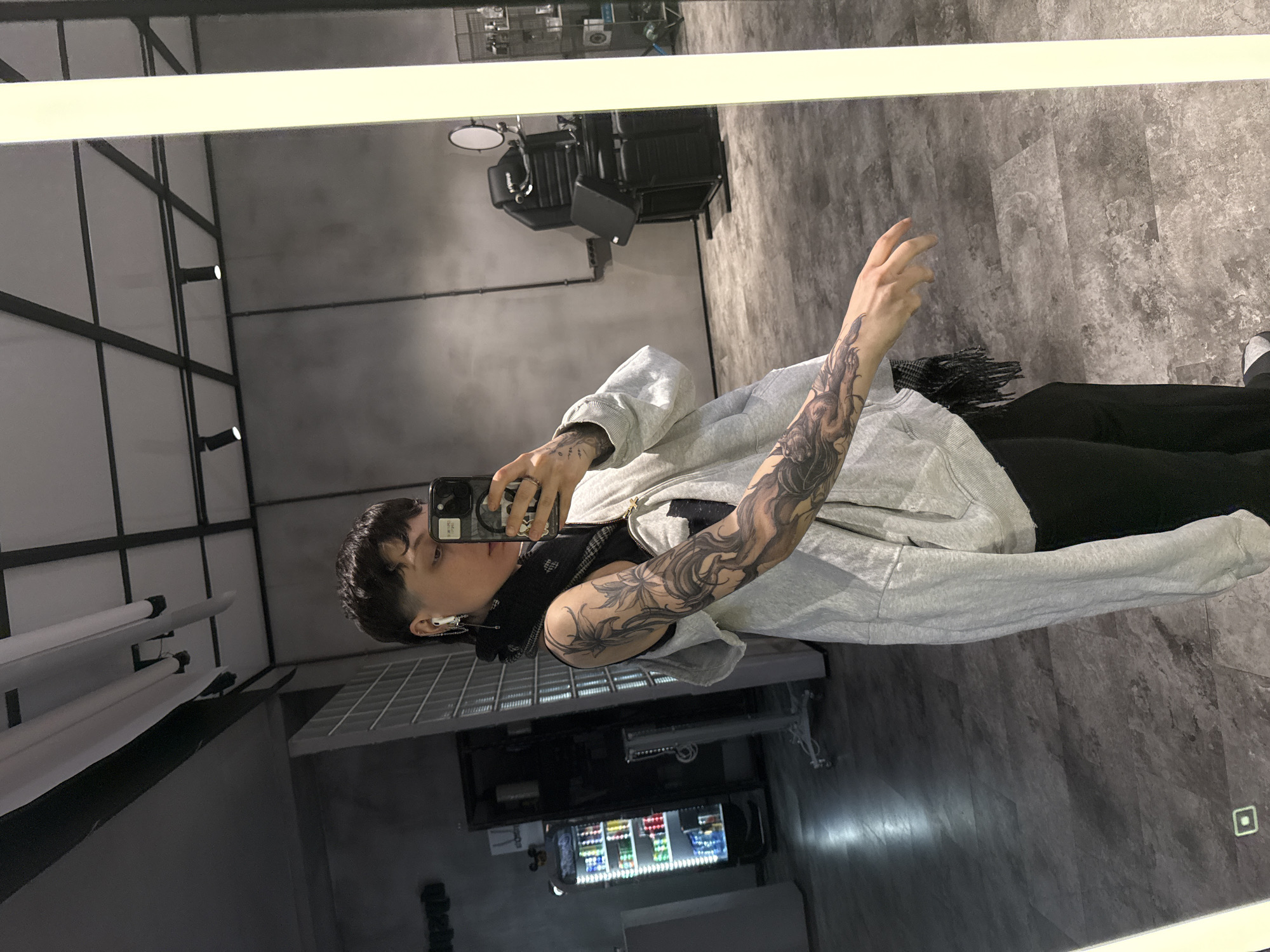 A person stands in front of a mirror, likely inside a business, judging by the barber's chair and cooler visible in the reflection. The individual, with short dark hair, wears a light grey hooded sweatshirt over a black undershirt, black pants, and what appears to be an earbud in their left ear. The person holds a smartphone up to take a selfie, their tattooed left arm prominently displayed.
The reflection shows a gray-toned interior. A barber's chair is visible, along with overhead lighting and some wall fixtures. A cooler is positioned to the side of the person's reflection. The location is unknown and no information in the image suggests this is in the city of Moscow, Russia.