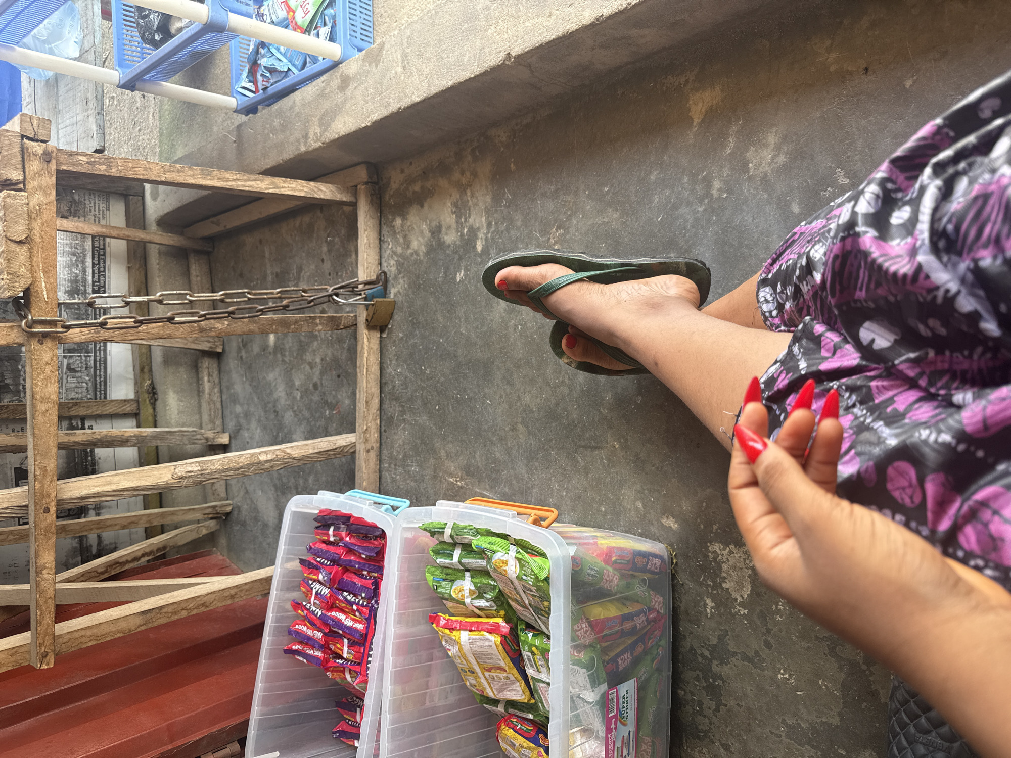 The image presents a ground-level view of an informal outdoor or semi-enclosed setting, likely in Uyo, Nigeria. A person is partially visible on the right side of the frame, with their legs extended. They are wearing a purple and black patterned garment and green flip-flops, with red nail polish visible on their toes. One hand, also featuring long, red-painted fingernails, is in the foreground.
In the lower center, two transparent plastic storage bins are filled with numerous individually packaged snacks or instant noodles. Text visible on these bins includes "SUPER PACK" and "SUPER STORER." On some of the snack packages, phrases like "Oodles Chicken" and what appears to be "BOOM SNACK" can be discerned.
To the left, a rustic wooden frame or structure, possibly part of a stall or fence, is prominent. A sturdy metal chain secured with a padlock is looped across one of its beams. Behind this wooden construction, fragments of newspaper or printed material are affixed, displaying text such as "Group Spring Up" and "OB Group." Above, a blue plastic shelving unit holds various items, including a black bag and plastic bottles. A red corrugated metal sheet is also visible on the lower left. The backdrop consists of a textured concrete wall and ground, indicating a utilitarian environment and suggesting a daytime setting. The scene implies a small vending operation or a storage area for goods.