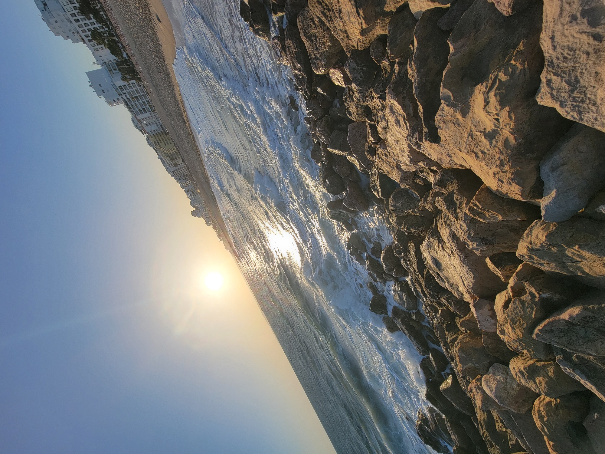 This is a rotated, eye-level, long shot of a rocky shoreline with the ocean and buildings. The sun is low in the sky, casting a warm glow and creating lens flare. The sky is clear and blue. Waves are crashing against a wall of large, rough-hewn rocks on the right side of the frame. To the left, there is a sandy beach, and beyond that, a line of white buildings that appear to be hotels or apartment complexes. The scene suggests a coastal town in Portugal, possibly Quarteira, during either sunrise or sunset. The overall mood is serene and picturesque. There is no visible text in the image.