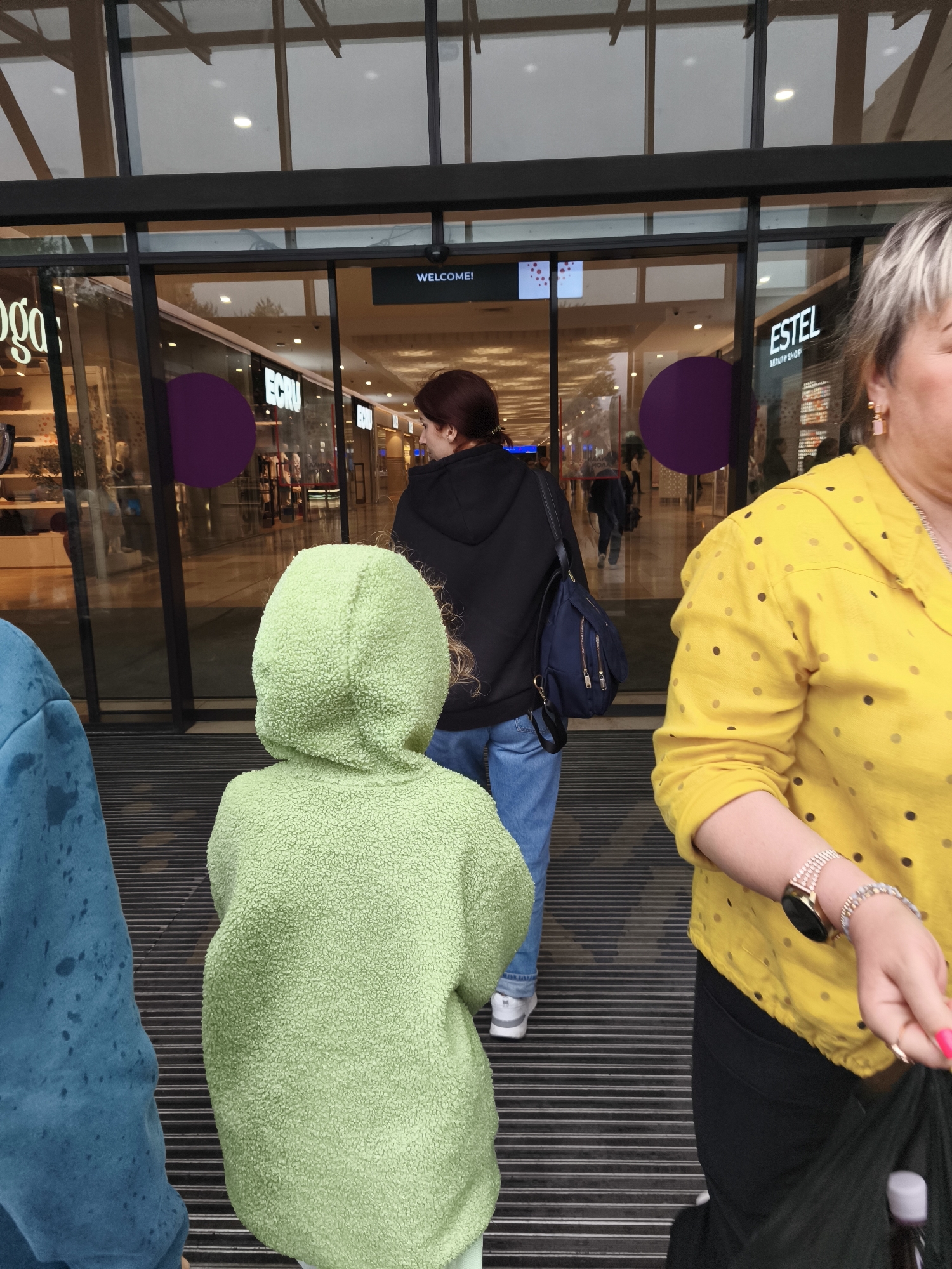 The image depicts the entrance to a shopping center with automatic glass doors. Inside, there are various storefronts, including one labeled "ECRU" and another with "ESTEL BEAUTY SHOP". Several people are visible. In the foreground, a person wearing a bright blue hooded jacket is on the left, partially cut off by the frame. Next to them, a child in a light green fuzzy hooded jacket is seen from behind, facing the entrance. To the right, a woman in a yellow polka-dot shirt and black pants is visible from the chest down, wearing a watch on her left wrist. Another woman in a black hoodie and blue jeans is walking away from the camera towards the inside of the shopping center. The ground in front of the entrance appears to be a striped walkway. The overall impression is a casual scene outside a commercial establishment.