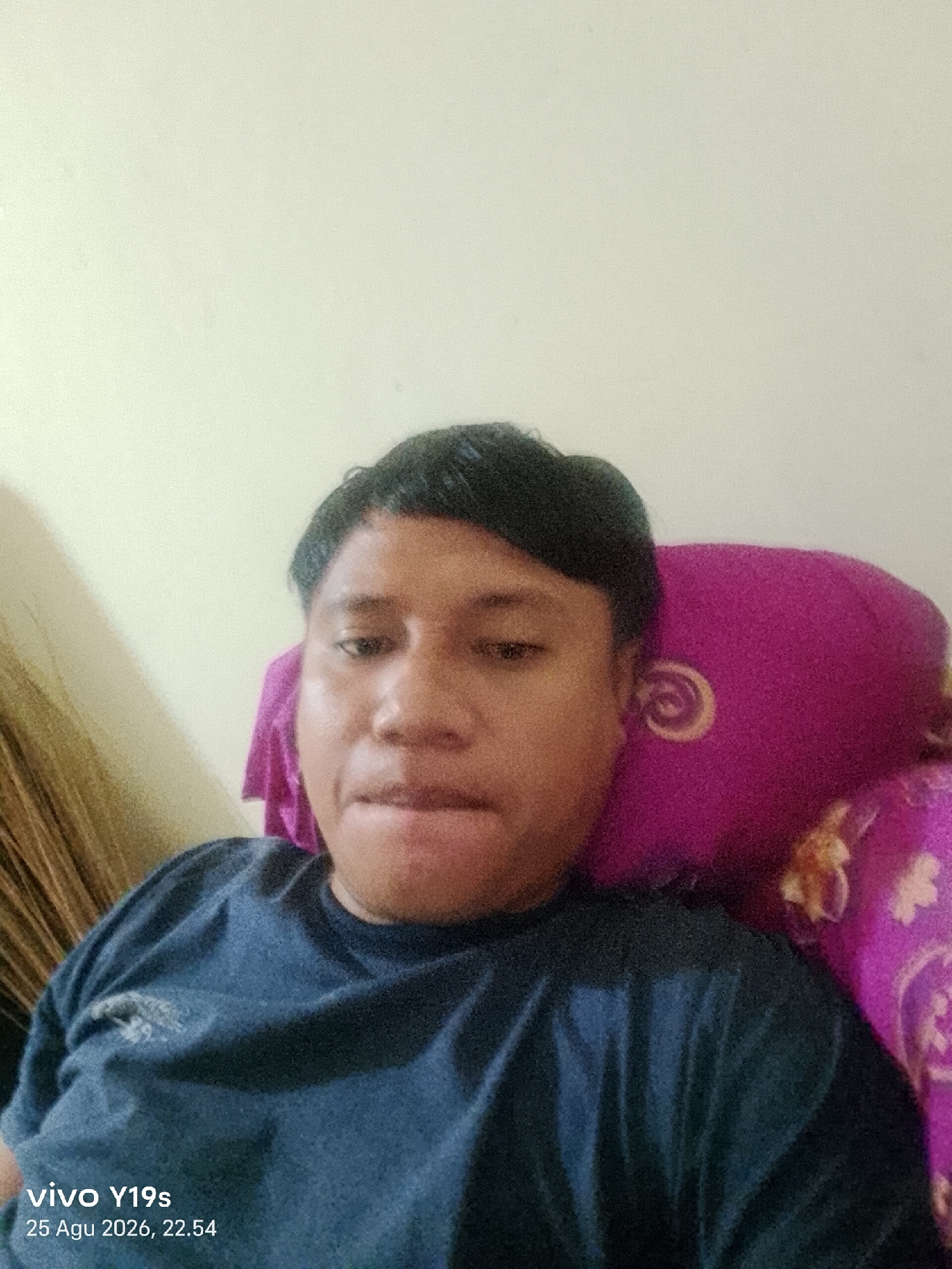 A selfie taken by a man in Ranoyapo, Indonesia, shows him from the chest up. He is wearing a dark blue t-shirt. A bright pink pillow with a swirl pattern is behind his right shoulder. The background is a plain, lightly colored wall. The bottom left corner of the image displays "vivo Y19s" and "25 Agu 2026, 22.54."