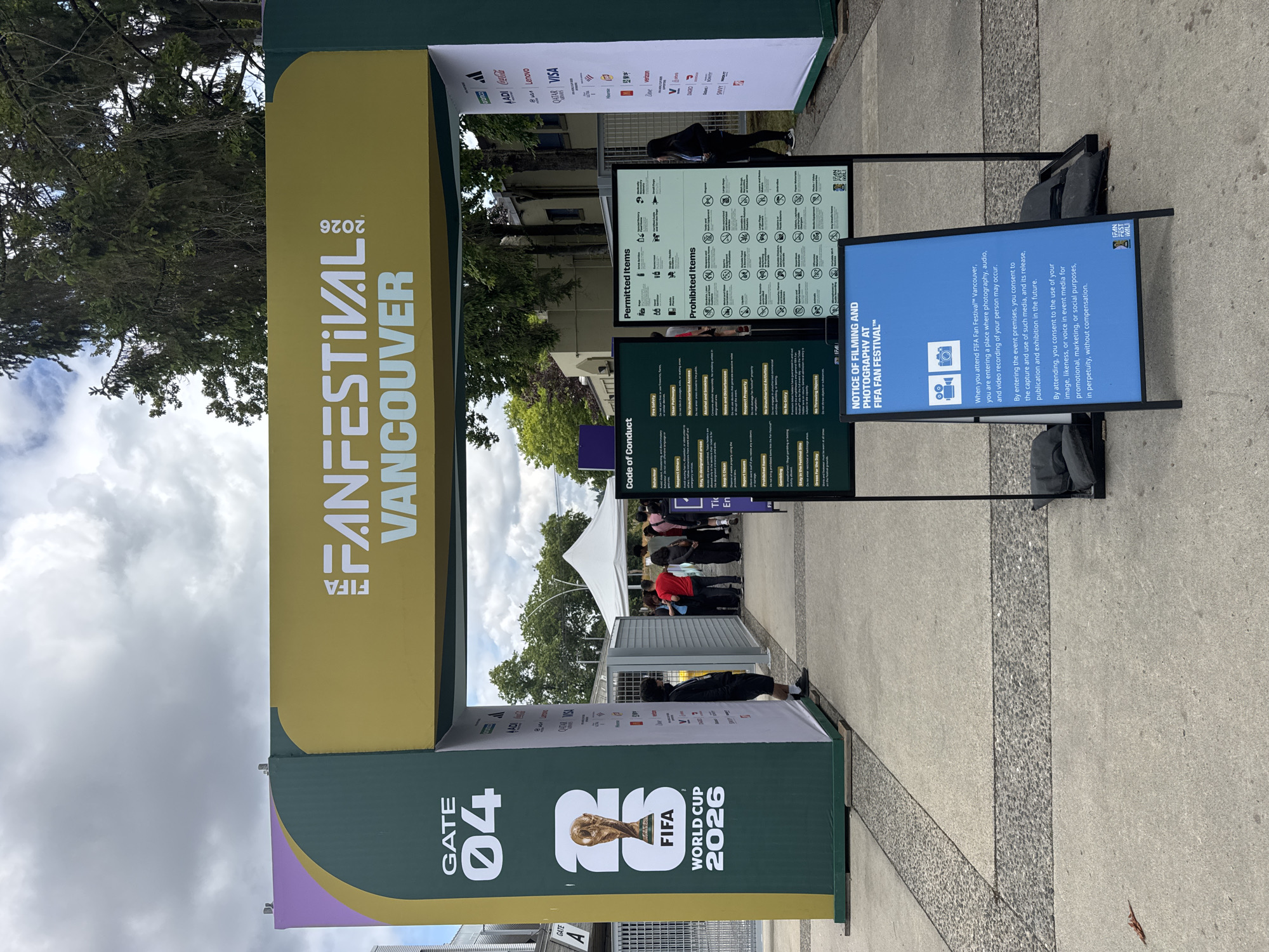 The image shows an entrance archway for the FIFA Fan Festival in Vancouver, Canada, with the year 2026 prominently displayed. The archway is decorated with the FIFA Fan Festival branding and features "GATE 04" on the left side, along with a graphic of the FIFA World Cup trophy. Several sponsors' logos are visible on the right side of the archway.
In front of the archway, there are two informational signs. The sign on the left details the "Code of Conduct" for attendees, outlining rules and regulations. The sign on the right lists "Permitted Items" and "Prohibited Items." A third, larger blue sign in the foreground, titled "NOTICE OF FILMING AND PHOTOGRAPHY AT FIFA FAN FESTIVAL™", explains the consent given by attendees regarding the capture and use of their image, audio, and video recordings.
Several people are visible in the background, queuing to enter the festival area, with a white tent structure visible further inside. The ground is paved with concrete and decorative gravel. Trees and buildings form the backdrop, under a partly cloudy sky.