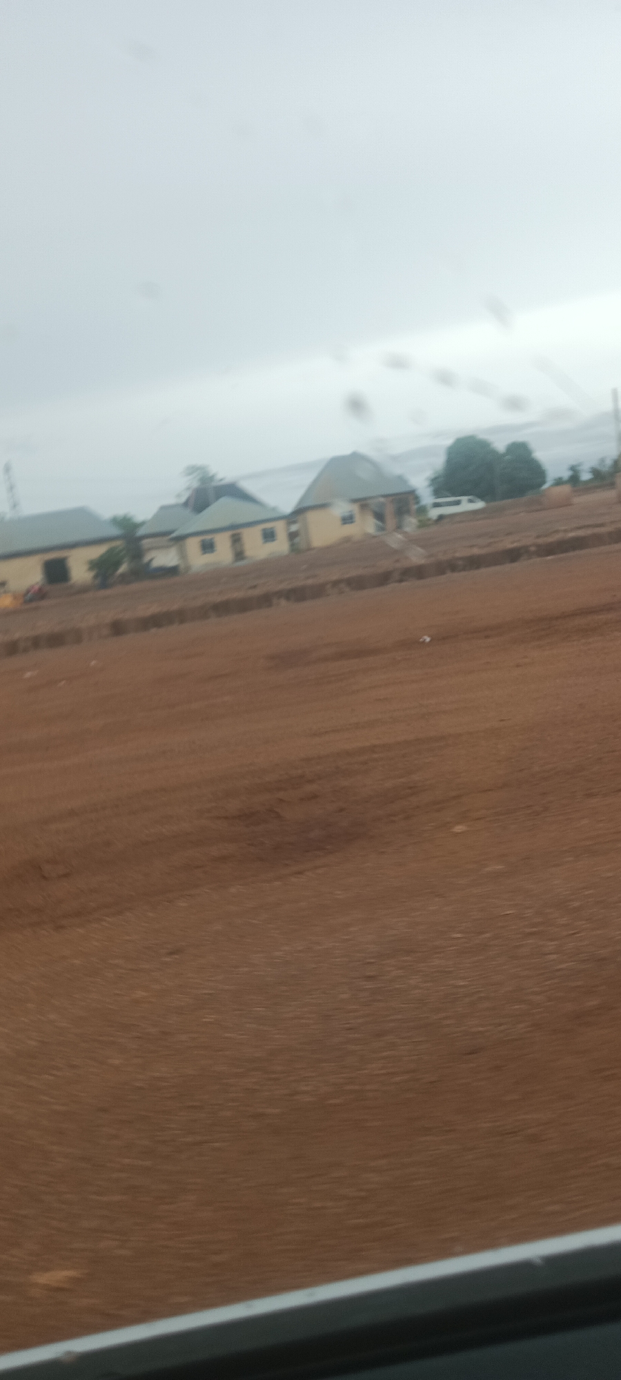 The image shows a roadside view in what appears to be Lafia, Nigeria. The foreground is dominated by reddish-brown soil. Several single-story buildings are visible in the midground, painted in a pale yellow color with grey roofs. A white van is parked further behind the buildings. The sky is overcast and grey. Power lines are visible in the background. There are no people visible in the image. The picture appears to have been taken from a moving vehicle, possibly a car, due to the motion blur effect on the foreground and the visible part of the vehicle's interior at the bottom of the frame.