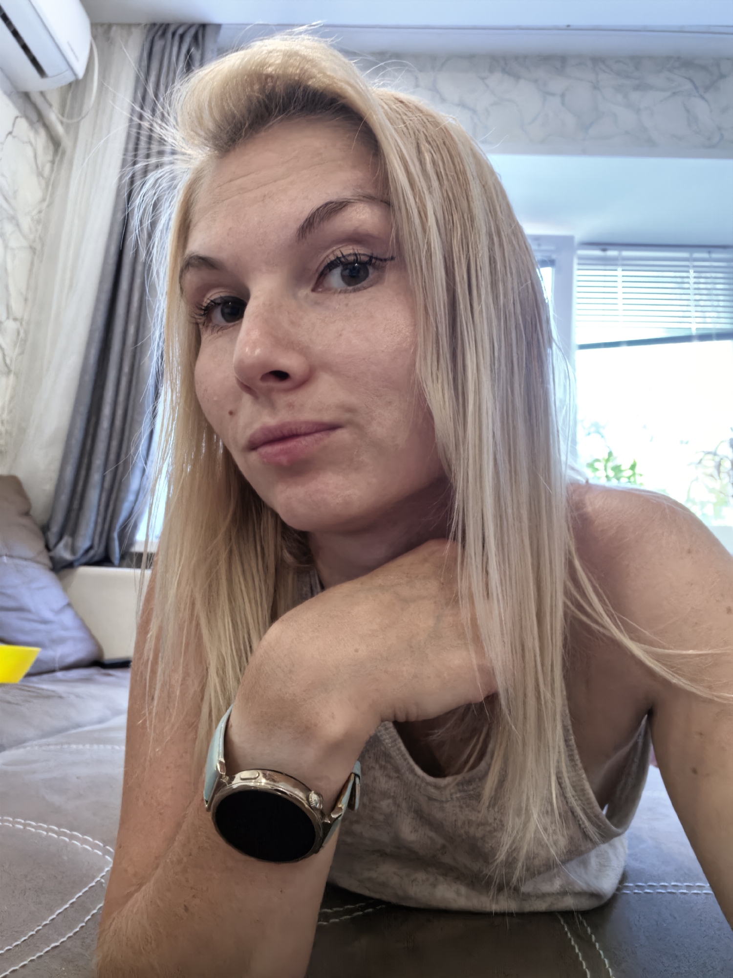 The image shows a medium shot of a woman with blonde hair, wearing a grey tank top and a smartwatch on her left wrist. She is leaning on her left arm, with her head tilted. Her gaze is directed towards the camera. The background appears to be an indoor setting with curtains, a window with blinds, and a couch. There is no information in the image that can be used to determine its location within the city of Sterlitamak, Russia.