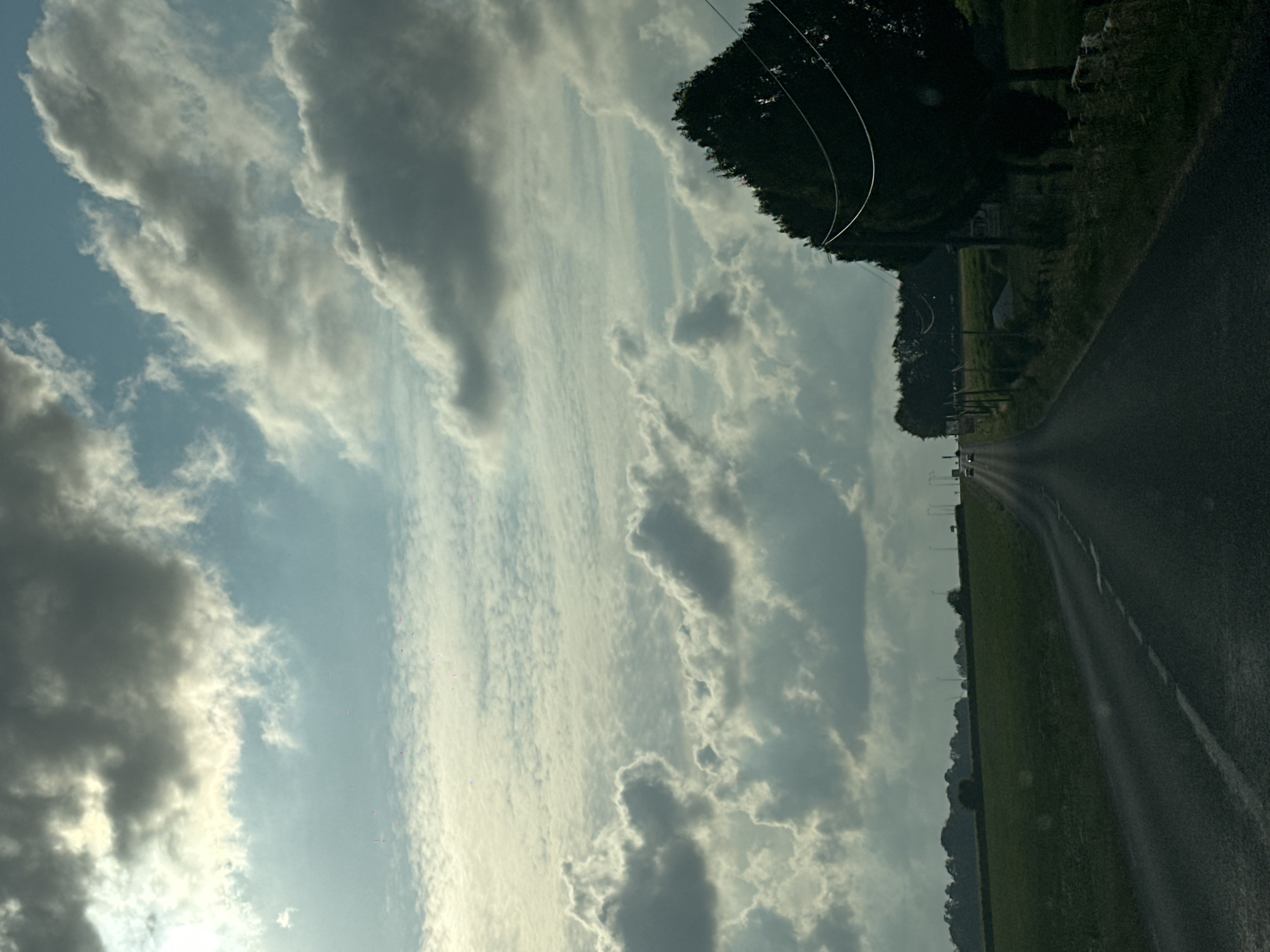 The image shows a view from a car on a road in the countryside, with a drone possibly in the distance or obscured by clouds. The sky is filled with dramatic, grey and white clouds, with patches of lighter sky and sunlight filtering through. A dark green tree, likely a conifer, is visible on the right side of the frame, with power lines strung across it. The road stretches into the distance, leading towards a line of trees and fields. The overall impression is one of a slightly overcast day in a rural setting. The location is stated to be in the city of Grigneuseville, France.