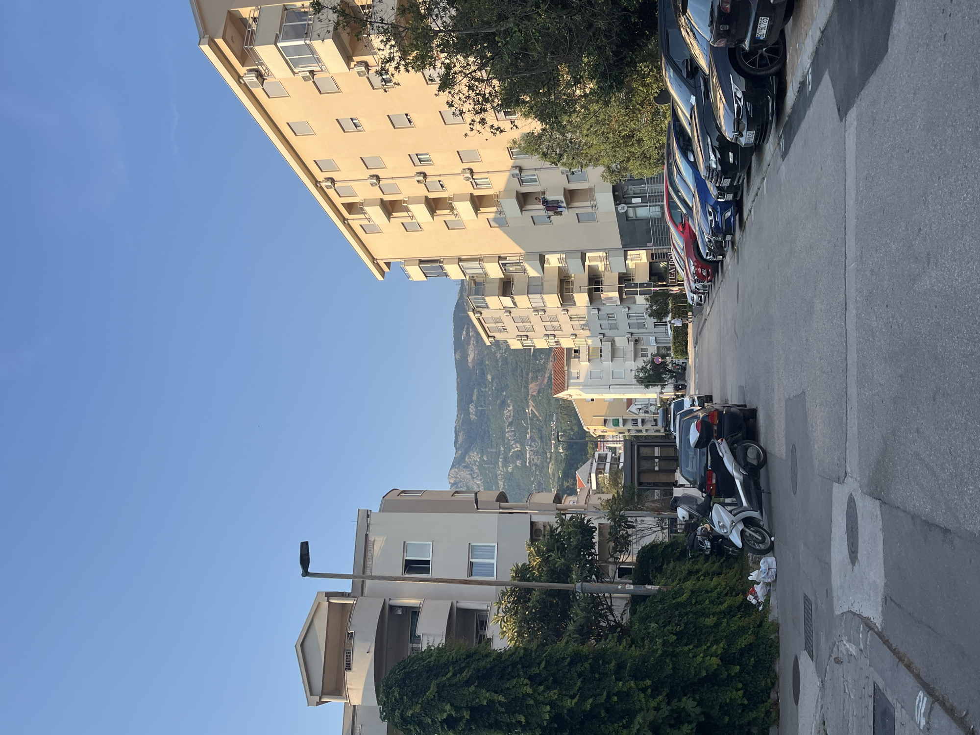 The image depicts a street scene in Budva, Montenegro, likely during the late afternoon or early evening, given the long shadows and warm, clear sky. The perspective is from a street level looking upwards and down a road lined with multi-story buildings and parked vehicles.
Dominating the left side of the frame are apartment buildings with cream-colored facades, featuring numerous windows and balconies. The architectural style suggests a Mediterranean influence. In the background, a majestic, verdant mountain rises, partially obscured by trees and other structures, characteristic of the Montenegrin landscape.
The street itself is paved, with a curb running along the side. Parked along the curb are several cars of various colors, including black, blue, and red. Two motorcycles, one silver and one white, are also parked. Litter is visible near the motorcycles.
Notable details include air conditioning units mounted on the buildings, and a street lamp is visible. Some windows are open, and on one balcony, two people can be seen, suggesting a residential area. The overall mood is peaceful and ordinary, capturing a slice of daily life in this coastal town. The clear blue sky indicates fair weather. No specific text is clearly legible except for what appears to be a sign for a "TEATRO" on one of the buildings, hinting at cultural amenities in the vicinity.