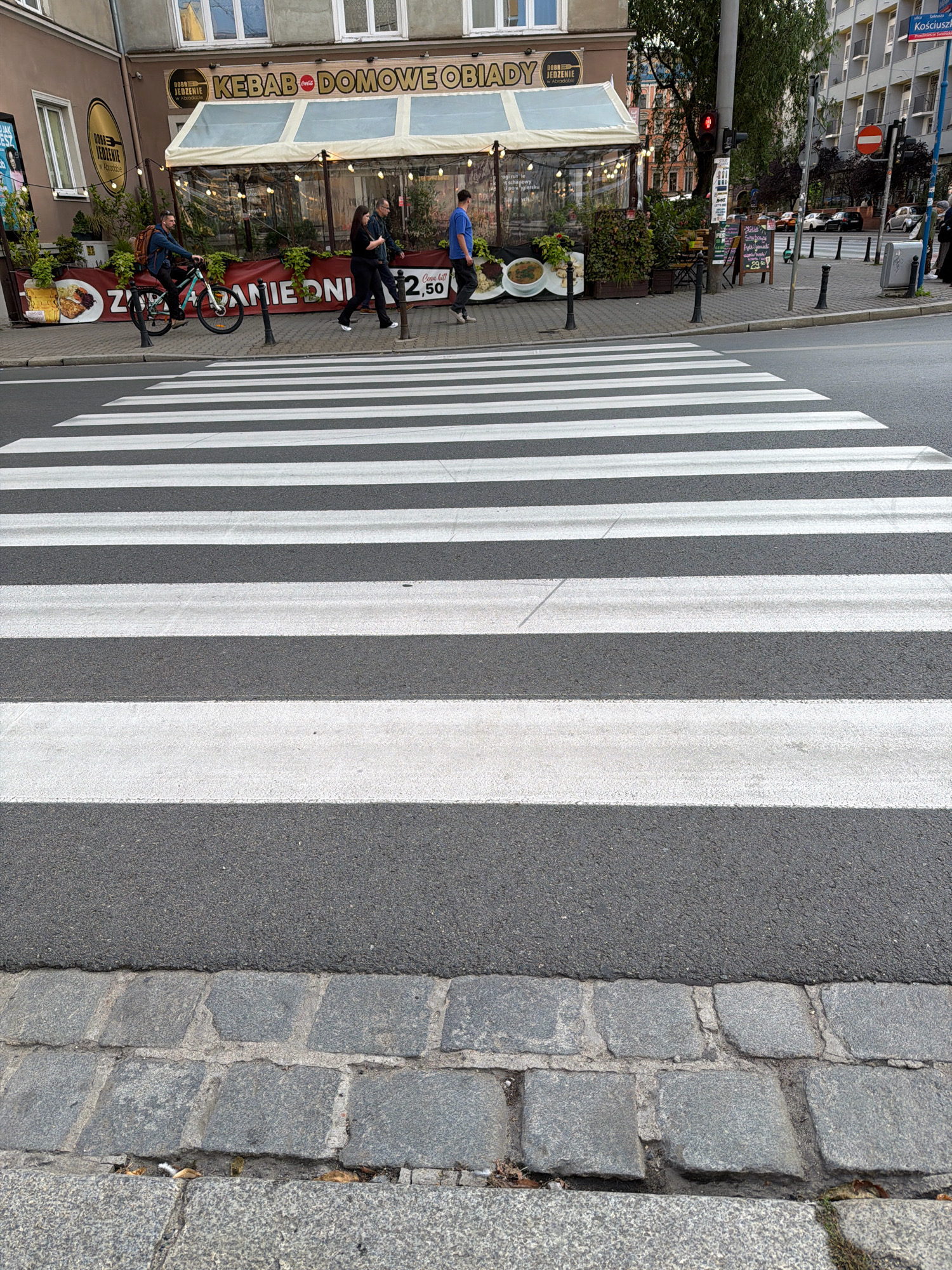 In Wrocław, Poland, a wide pedestrian crosswalk dominates the foreground, its white stripes stark against the asphalt. The crosswalk is bordered by cobblestone paving. Beyond the crosswalk, a street scene unfolds. A man on a bicycle, wearing a backpack, rides past a restaurant with a covered outdoor seating area. The restaurant displays a banner that reads "KEBAB DOMOWE OBIADY." Several people are walking on the sidewalk, including a couple and another man. In the background, buildings of varying architectural styles line the street, and traffic lights and street signs are visible. A red traffic light is illuminated, indicating that vehicles must stop. The overall impression is of a bustling urban environment.