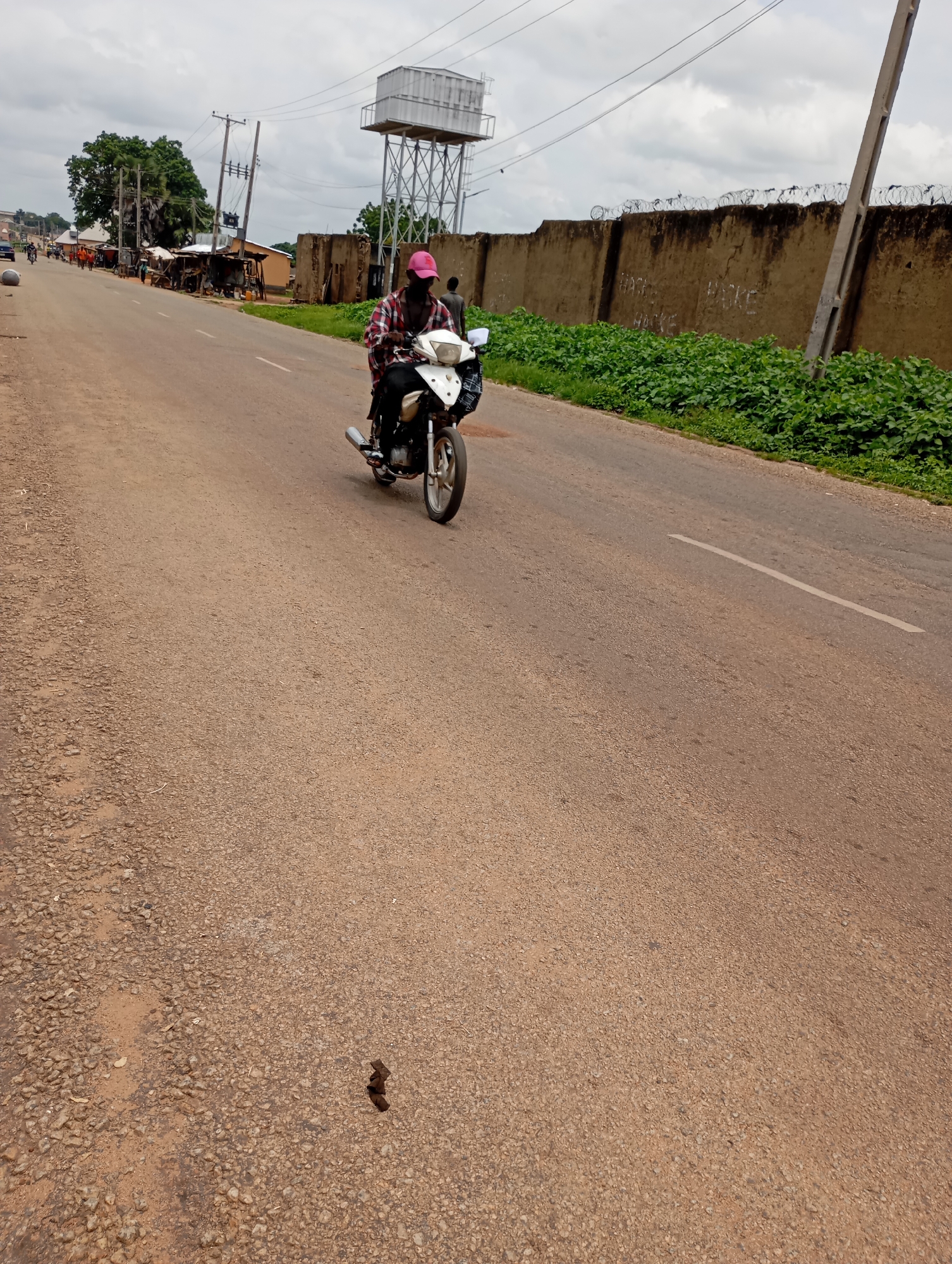 The image depicts a street scene in Shendam, Nigeria. A person wearing a pink cap and a red and white checkered shirt rides a white motorcycle on a dusty, paved road. The motorcycle has a silver exhaust pipe and a black rear basket. On the left side of the road, there are small shops with awnings and some trees. In the background, a tall water tower on a metal frame is visible, along with power lines and poles. A concrete wall with barbed wire on top runs along the right side of the road. Green vegetation grows along the base of the wall. The sky is overcast with clouds. There are some scattered objects and debris on the road, including what appears to be animal droppings in the lower center of the image.