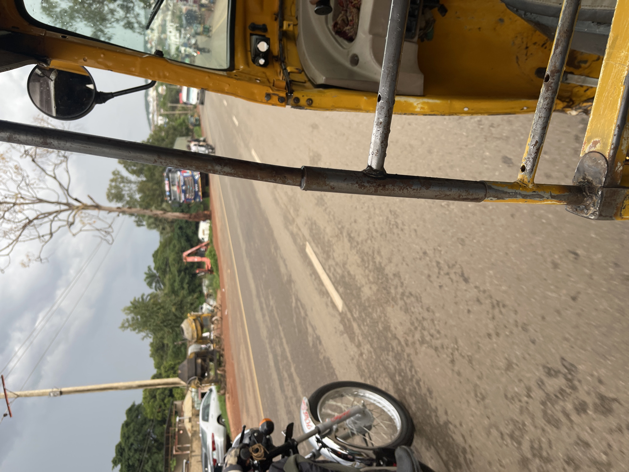 A street-level, medium-distance, eye-level shot shows a yellow auto-rickshaw moving on a paved road. The camera is inside the auto-rickshaw, focused on the passenger side. The road is wet, with water stains and streaks from recent rain. The sky is overcast with a greyish hue. On the left side of the road, there are several parked vehicles, including a white sedan and a motorcycle. In the background, there are construction materials, including two yellow cement mixers and red construction equipment. Further in the distance, a large blue truck with red accents is visible, along with other vehicles on the road and a hillside with buildings. The environment appears to be a semi-urban area, with trees and vegetation lining the road. The overall scene depicts a typical day on the road in Rafin Par, Nigeria, characterized by the presence of auto-rickshaws, local traffic, and ongoing construction.