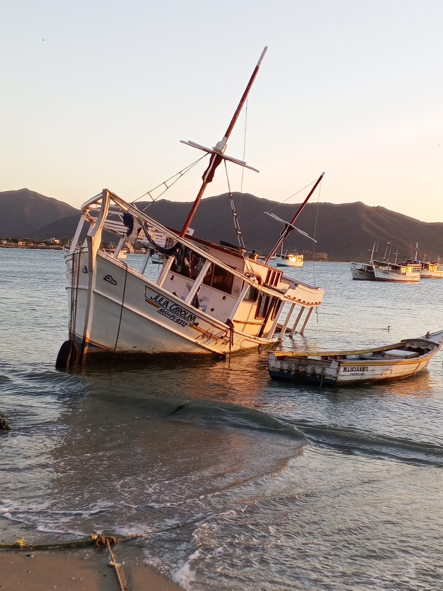 The image shows a damaged, white fishing boat, identified as "Julia Carolina ARSH-PE 0312", listing severely to one side in shallow water near a beach. The boat is partially submerged, and its structure appears weathered and damaged. Alongside it is a smaller boat, "M.Luciannis Fanilova," also showing signs of wear. In the background, the landscape reveals a body of water with several other boats anchored or moored further out. Mountains or hills form the backdrop, and the sky suggests either sunrise or sunset, casting a warm light across the scene. The location is Juan Griego, Venezuela, as indicated by the boat's registration. The beach and water show evidence of human presence and boat activity, but no individuals are directly visible in the frame.