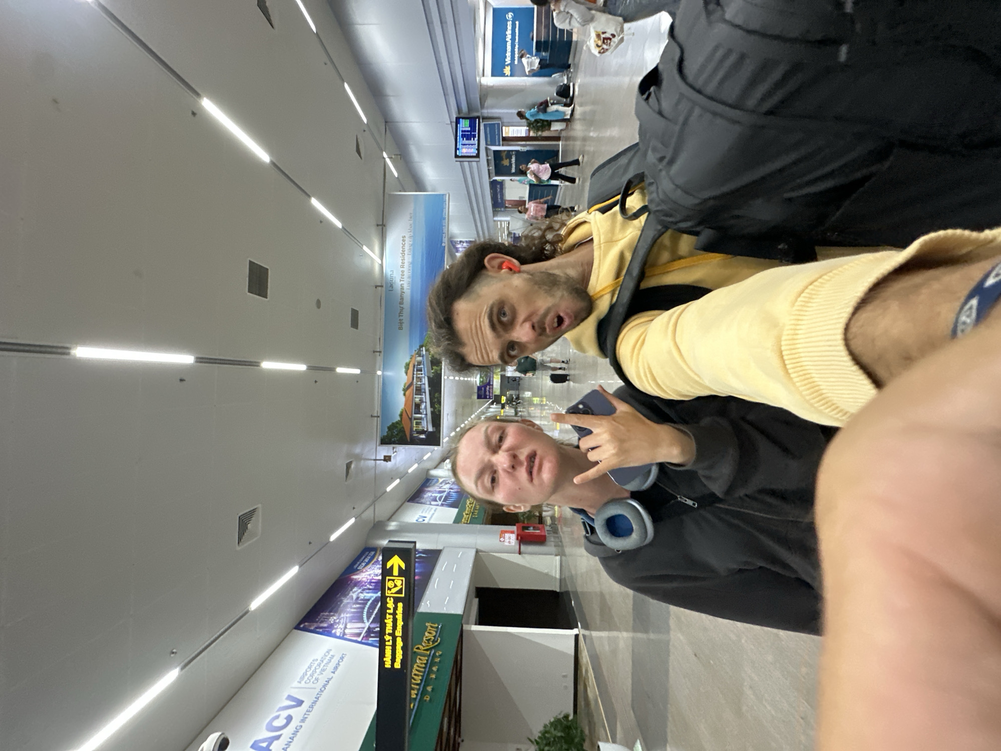 This is a medium shot, taken from a low angle, of a couple in an airport terminal. The person on the left, a woman with blonde hair, is making a rock-on hand gesture and sticking out her tongue at the camera, while holding a phone in her other hand. She is wearing a dark grey hooded jacket with blue headphones around her neck. The person on the right, a man with dark, curly hair, is making a surprised expression with his mouth open, and is wearing a yellow hoodie. He has red earbuds in both ears and is carrying a black backpack. The background of the image shows an airport interior with a tiled floor and a high ceiling with fluorescent lighting. There are signs in Vietnamese and English, one of which indicates "Baggage Enquiries" with a yellow arrow pointing to the right, suggesting the location is Da Nang International Airport in Da Nang, Vietnam. Other signs and people in the background indicate a typical airport environment with check-in counters and departure boards. The image appears to be a selfie.