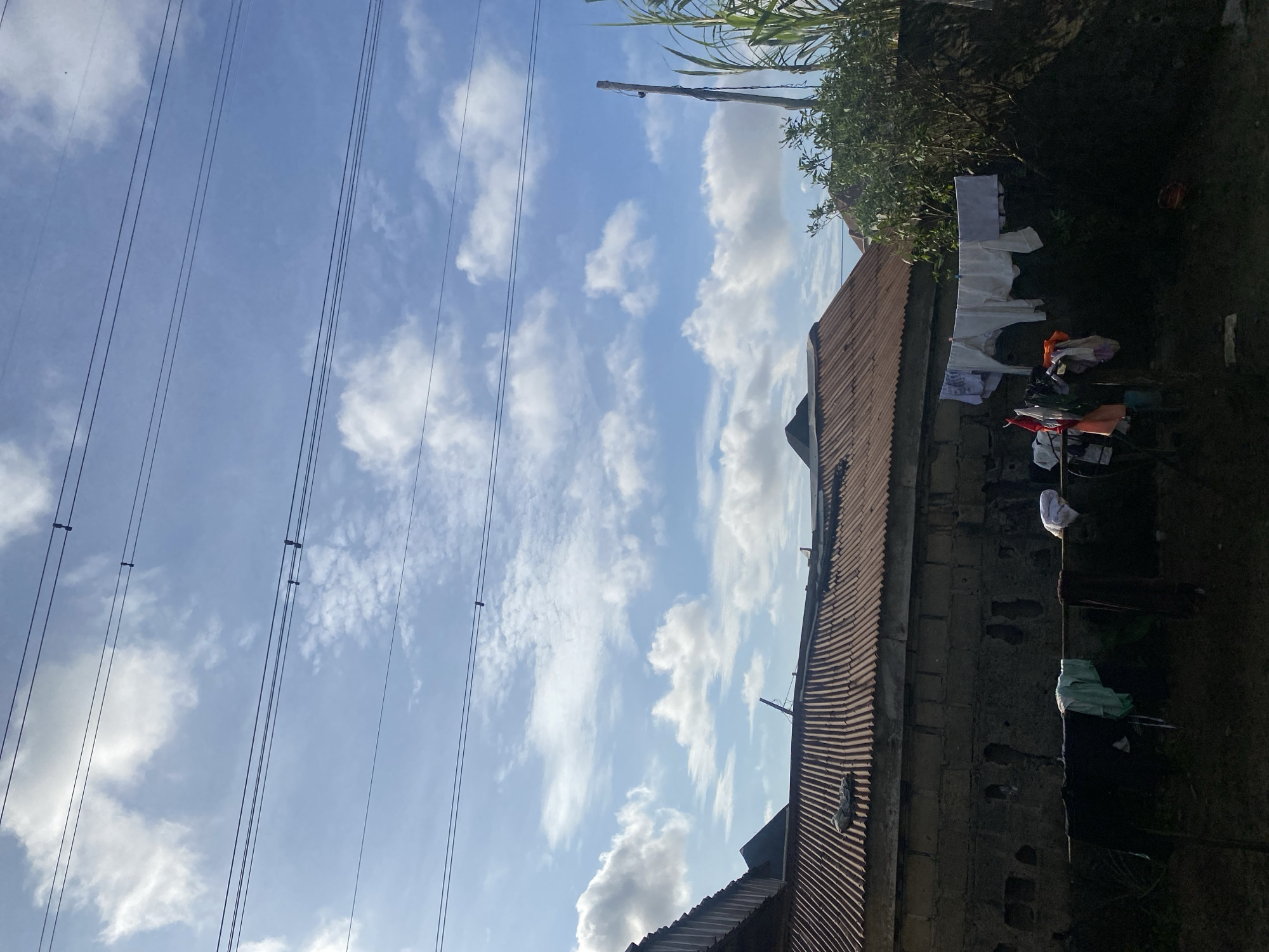 This is an eye-level, medium-distance, outdoor shot of a residential area, likely in Ado, Nigeria, under a partly cloudy sky. The scene is dominated by a rustic, corrugated iron roof of a building, possibly made of cinder blocks. In the foreground, clothes are hung out to dry on clotheslines, with a mix of light-colored garments and darker items. A wooden pole with what appears to be a camera or sensor is visible on the right side of the image, partially obscured by lush green foliage. Power lines crisscross the sky. The overall impression is one of everyday life in a developing urban environment.