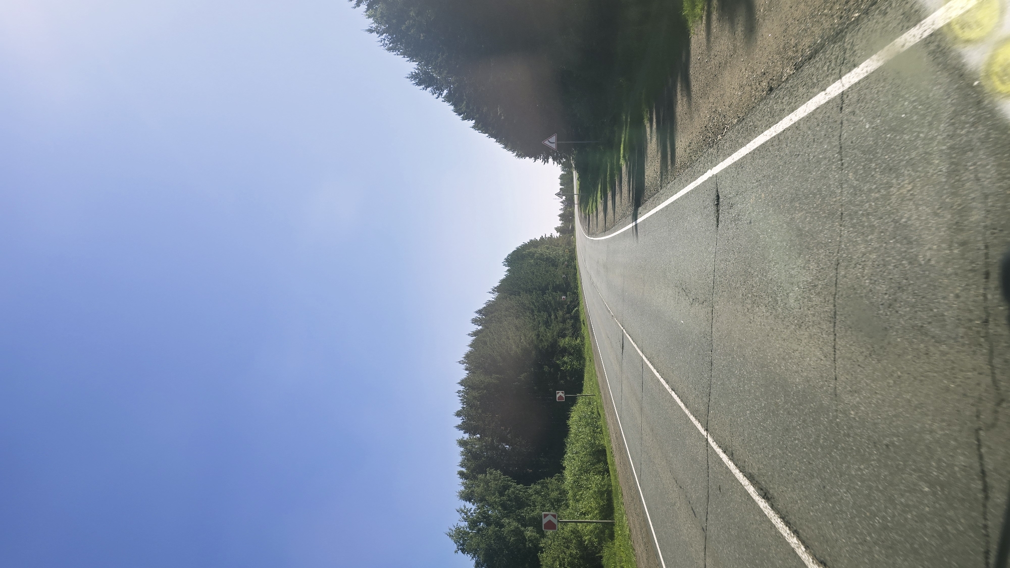 The image depicts a two-lane asphalt road curving to the right, bordered by dense green trees on both sides. The sky above is a clear, pale blue. On the left side of the road, there are signs with red circular markers, indicating a turn. Further down the road, on the right side, there is a triangular warning sign. The road surface shows signs of wear with visible cracks. The scene appears to be in a rural or forested area, possibly on the outskirts of a city. The lighting suggests it is daytime, with the sun casting shadows from the trees onto the road. The specific location in the city of Актюбинский, Russia cannot be determined solely from the visual information in the image.