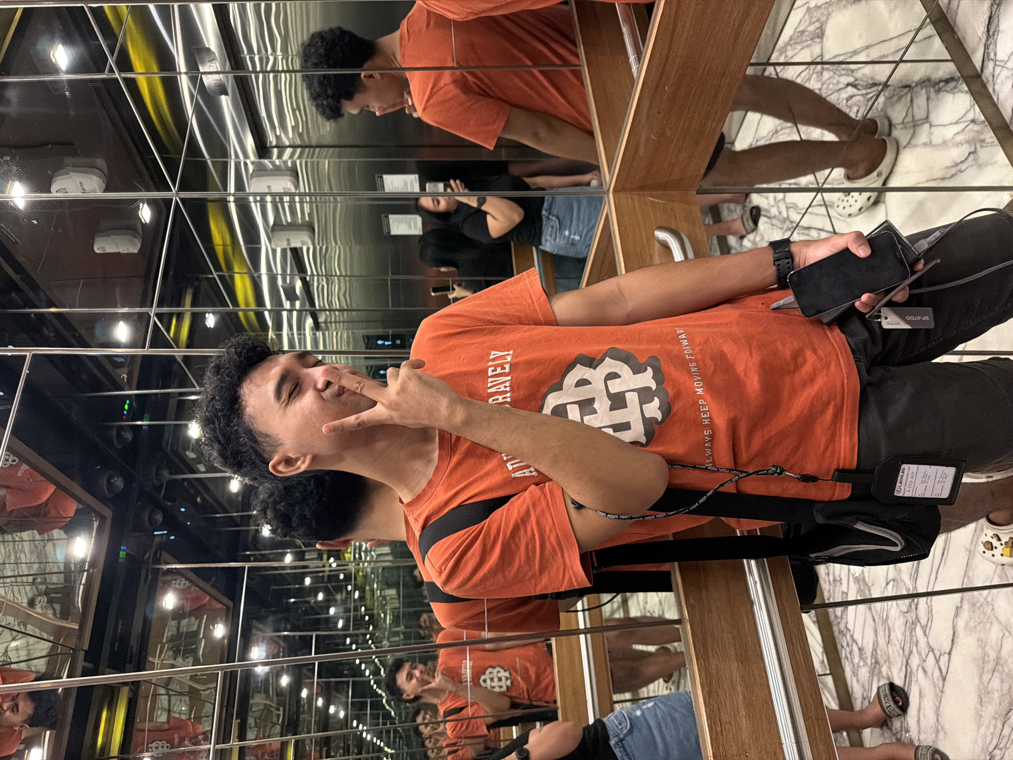 In the mirrored interior of an elevator, a young man with curly dark hair, wearing an orange t-shirt with a graphic logo and dark shorts, poses with his eyes closed and his fingers near his mouth, making a peace sign. He holds a smartphone and a portable charger in his right hand. A black bag with a lanyard is slung over his shoulder. The elevator walls are lined with mirrors, reflecting the man, other people, and the elevator's interior, including metal doors, lighting fixtures, and a wooden railing. On the marble-patterned floor, various individuals are visible in the reflections, including a woman in a black top and denim shorts holding a phone, and another person wearing sandals. The elevator appears to be in a commercial building, possibly a mall or hotel, located in Central Jakarta, Indonesia.