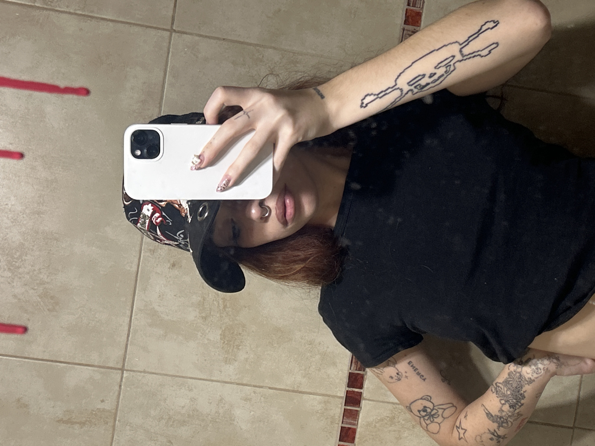 A woman takes a selfie in front of a mirror. The subject is wearing a black t-shirt and a baseball cap with a black bill and decorative trim. Her right arm, visible in the upper right corner, bears a tattoo. The phone in her hand is white. Her hair is reddish-brown. She has a nose ring. Tattoos are visible on her left arm as well. The backdrop consists of tiled flooring and a tiled wall. The scene is not in the city of Buenos Aires, Argentina.