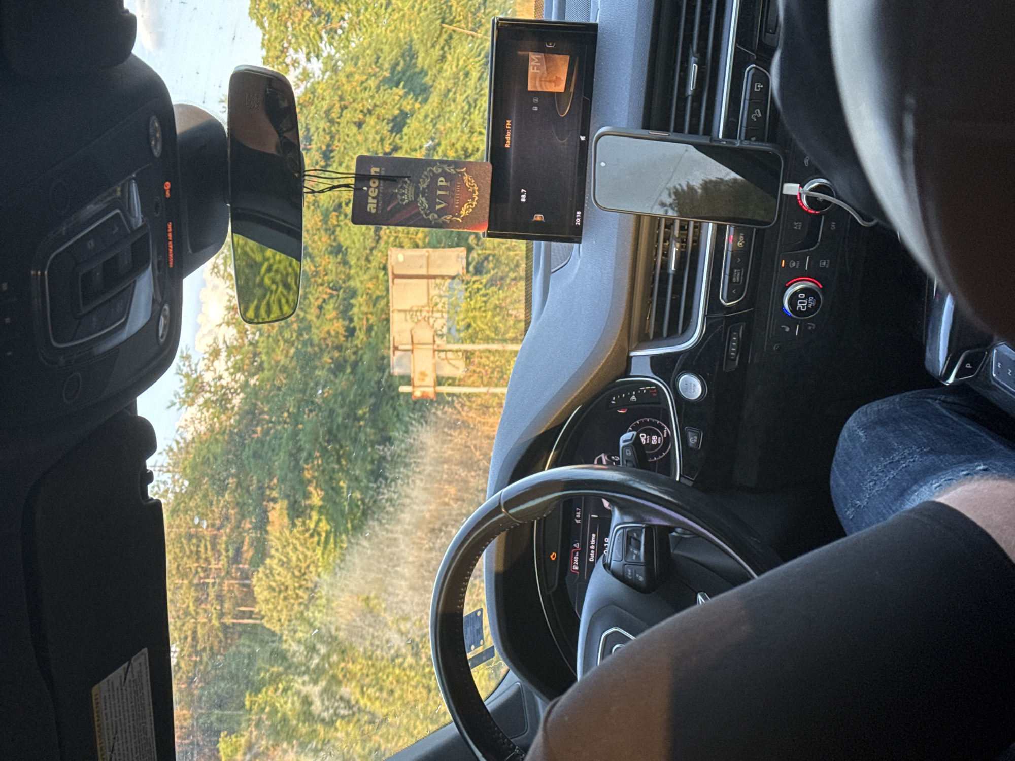 The image presents the interior of an Audi Q7 vehicle, observed from the front passenger seat, situated in Strumyani, Bulgaria. Through the windshield, a dense forest bathed in golden hour light is visible, with a faded brown road sign partially discernible in the mid-ground. The driver is seated, with their left arm in a black t-shirt and legs in blue distressed jeans visible. The steering wheel, featuring the Audi logo, is in the foreground. The instrument cluster displays a range of 240 km, a radio frequency of 88.7, and a speed of 55 km/h, alongside an illuminated warning light (resembling a check engine icon). The central infotainment screen shows "Radio: FM", "88.7", and the time "20:10". An "Areon VIP EXCLUSIVE" air freshener hangs from the rearview mirror. Above the rearview mirror, the overhead console indicates "PASSENGER AIR BAG ON" in red. A smartphone is mounted vertically on the dashboard, actively charging via a white cable. Below, the climate control panel is set to "20.0 AUTO". The windshield exhibits minor specks and dust.