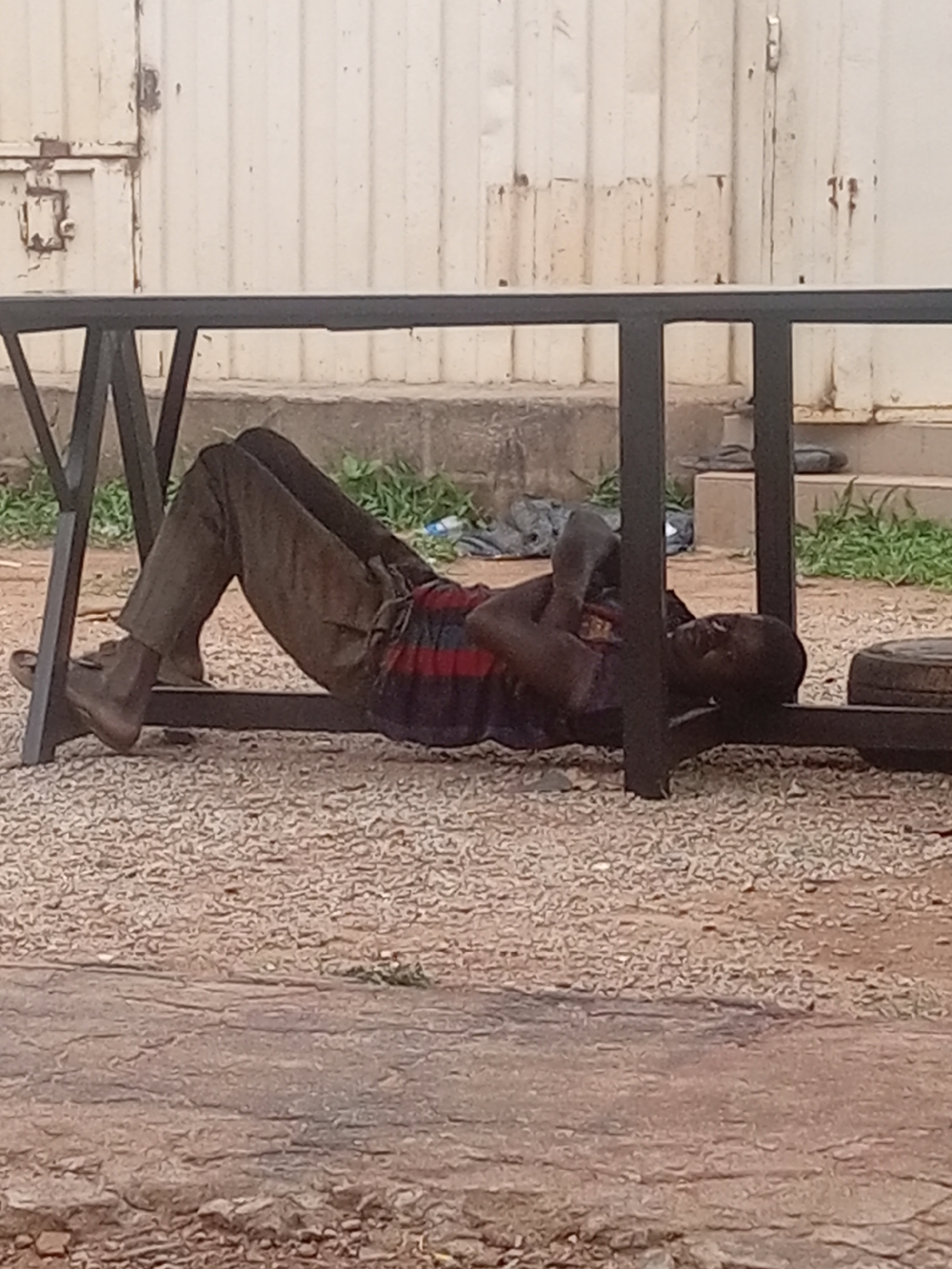 In the city of Unknown, Unknown, an adult male is observed lying supine on a light brown gravel surface, positioned beneath a dark, heavy-gauge metal frame structure. His head is oriented towards the right side of the frame, and his lower body towards the left. His legs are bent at the knees, with his feet, clad in brown shoes or sandals, flat on the ground. He is wearing dark trousers and a horizontally striped shirt featuring alternating dark blue/purple and red/maroon bands. His right arm is bent with the hand raised near his chest, possibly holding a small, dark rectangular object. His left arm is bent, resting across his torso. His eyes are open and directed forward. Behind the man and the metal frame, patches of green vegetation are visible, bordering a concrete curb. Beyond the curb, a tall, light-colored corrugated metal wall or building facade extends across the background, displaying evidence of rust and a visible latch mechanism on the left side. To the right of the man's head, partially obscured by the metal frame, a dark, circular object resembling a tire or a heavy weight rests on the gravel. The scene appears to be outdoors under daylight conditions.