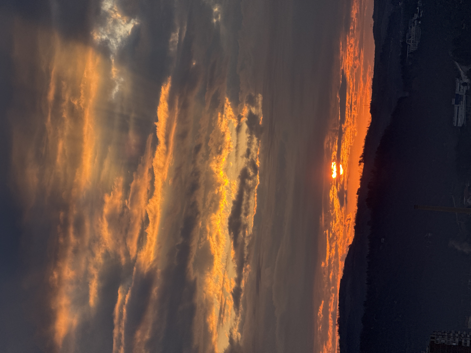The image captures a sunset in Veliko Tarnovo, Bulgaria. The sun, a vibrant orange, dips below the horizon along the silhouette of a forested hill. Wispy clouds, illuminated in shades of orange and gold by the setting sun, stretch across the sky. The foreground is dark, with faint outlines of buildings and structures, indicating the city's layout.