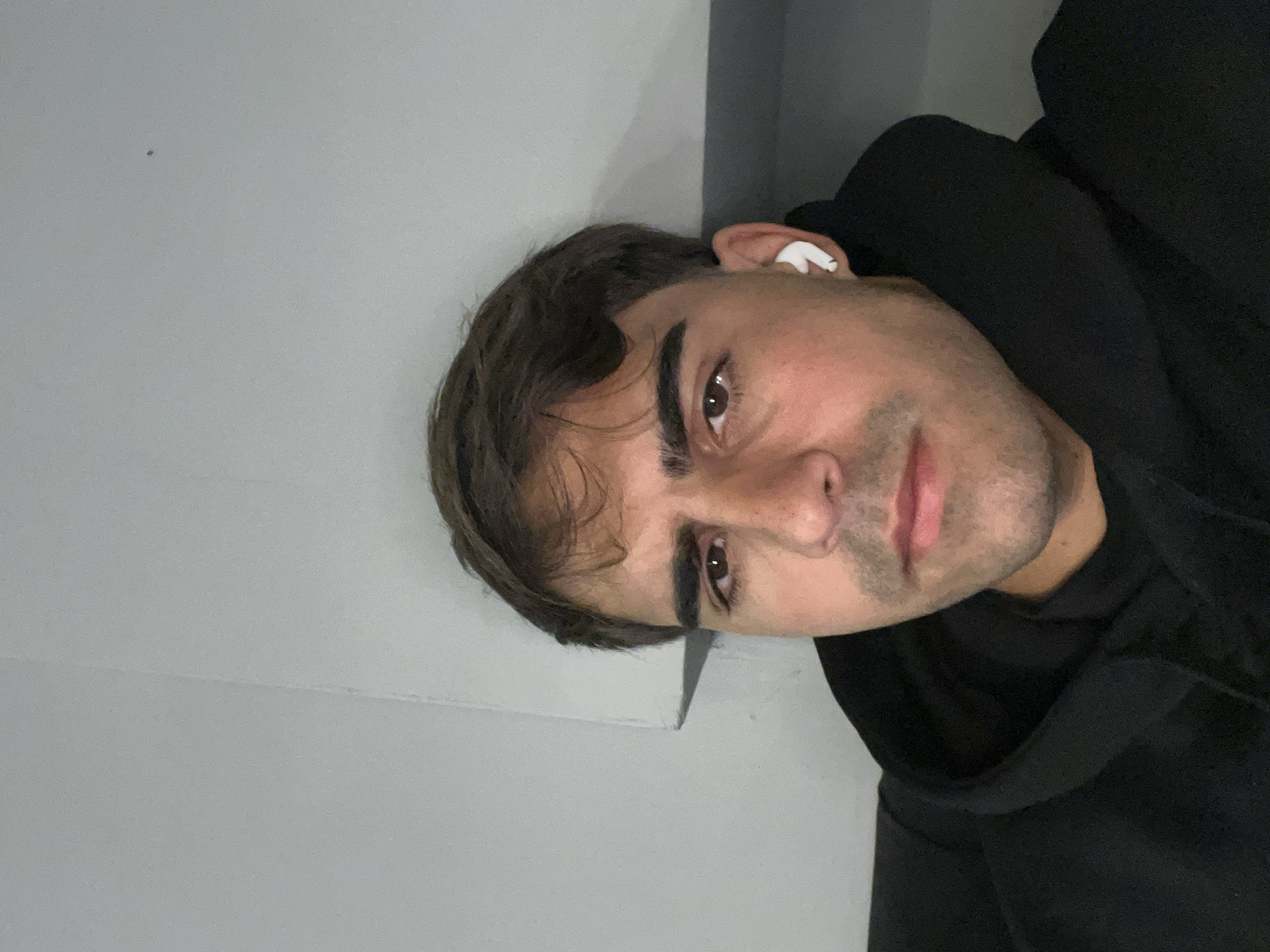 A young man, likely in his 20s or 30s, is featured in a close-up, head-and-shoulders portrait. The image is originally rotated 90 degrees counter-clockwise, but when viewed upright, he looks directly towards the viewer with a neutral, slightly contemplative expression. He has short, dark brown hair, which is slightly wavy and falls across his forehead. His eyes are dark brown and focused. He has well-defined eyebrows and light stubble on his chin and jawline. His lips are slightly parted.
The man is wearing a dark, possibly black, hooded sweatshirt or jacket, visible around his neck and shoulders. A white wireless earbud is clearly visible in his right ear. The background is a plain, light gray wall or ceiling, indicating an indoor setting. There's a subtle seam or corner visible in the upper left portion of the background when the image is viewed upright. The lighting appears soft and even, typical of indoor artificial light. No specific activity is taking place other than posing for the photograph. The location is indoors, within Buenos Aires, Argentina. There is no visible text in the image.