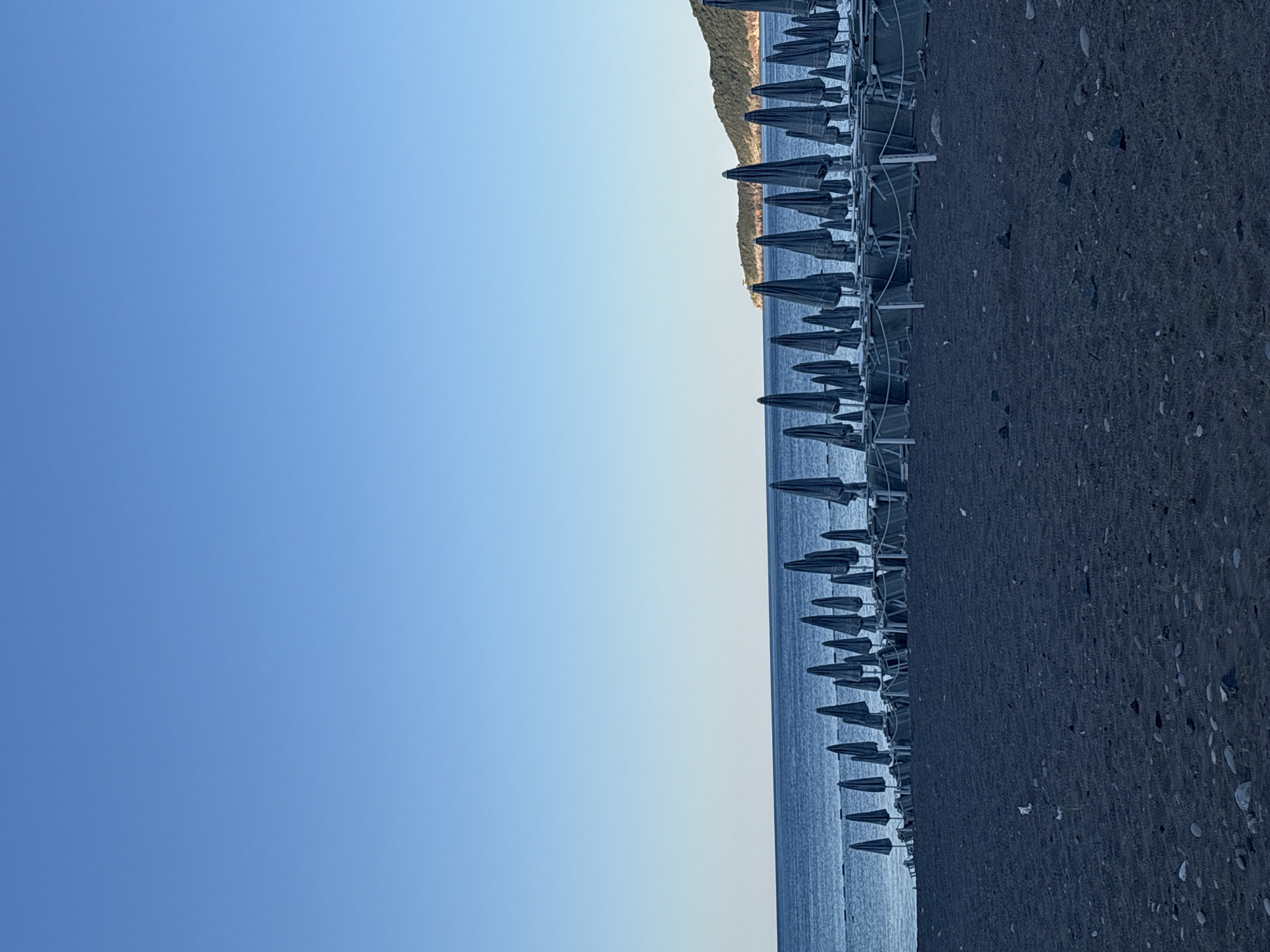 A vertical, low-angle shot captures a quiet beach scene in Bečići, Montenegro, under a clear blue sky. The foreground is dominated by dark, coarse sand interspersed with small pebbles. Behind the sand, a long row of closed beach umbrellas stands in neat rows, casting dark shadows. These umbrellas are dark blue or grey and are folded up on their poles, suggesting they are not in use. Behind the umbrellas, the calm, blue sea stretches to the horizon, meeting the sky in a sharp line. In the distance, a lush, green hillside with a rocky outline rises from the sea, a small white structure, possibly a lighthouse, is visible on its slope. The overall impression is one of tranquility and early morning or late evening stillness on the Adriatic coast.