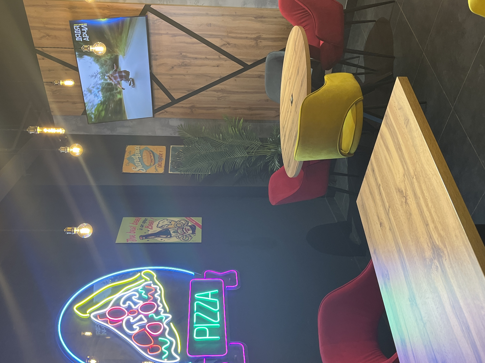 The image captures an interior scene in what appears to be a pizza restaurant in Izhevsk, Russia, judging from the Cyrillic script visible on the TV screen ("ДЯДЯ АРЧИ"). The restaurant features a mix of decor elements, including neon signs depicting a pizza slice and the word "PIZZA," vintage-style signs promoting sandwiches and beer, and string lights with Edison bulbs. There's also a TV screen playing a video. The furniture includes wooden tables of both round and rectangular shapes, surrounded by red and yellow chairs. A potted plant sits near one of the tables. The restaurant space has a dark color scheme, with dark walls and flooring.