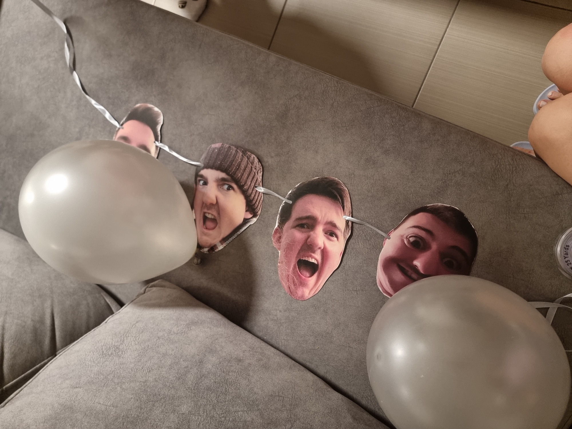 The image shows a close-up view of a grey couch with a string of paper cutouts of human faces attached to it. There are two silver balloons, one near the top of the image and another near the bottom left. The faces are printed on paper and appear to be attached to the string with tape or glue. The couch is in an indoor setting. The image does not provide any information about the location in the city of Ramat Gan, Israel.