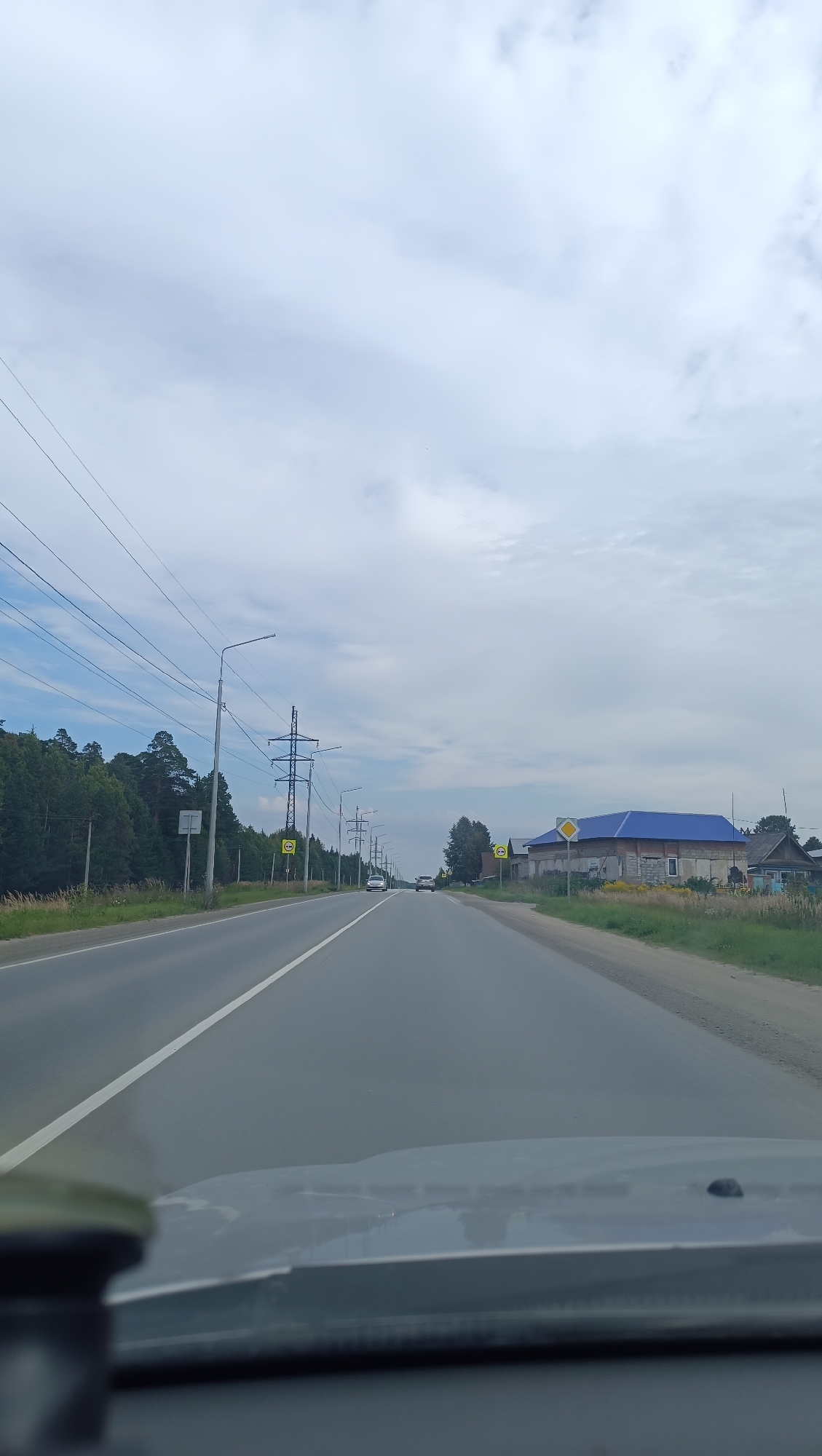 An outdoor, eye-level, wide shot shows a view from a vehicle traveling on a two-lane asphalt road under a cloudy sky. The road has a white line dividing the lanes. Trees and power lines line the left side of the road, while houses and a large building with a blue roof are visible on the right. Several cars are traveling in the same direction as the viewer. The foreground shows the dashboard and windshield of the vehicle.