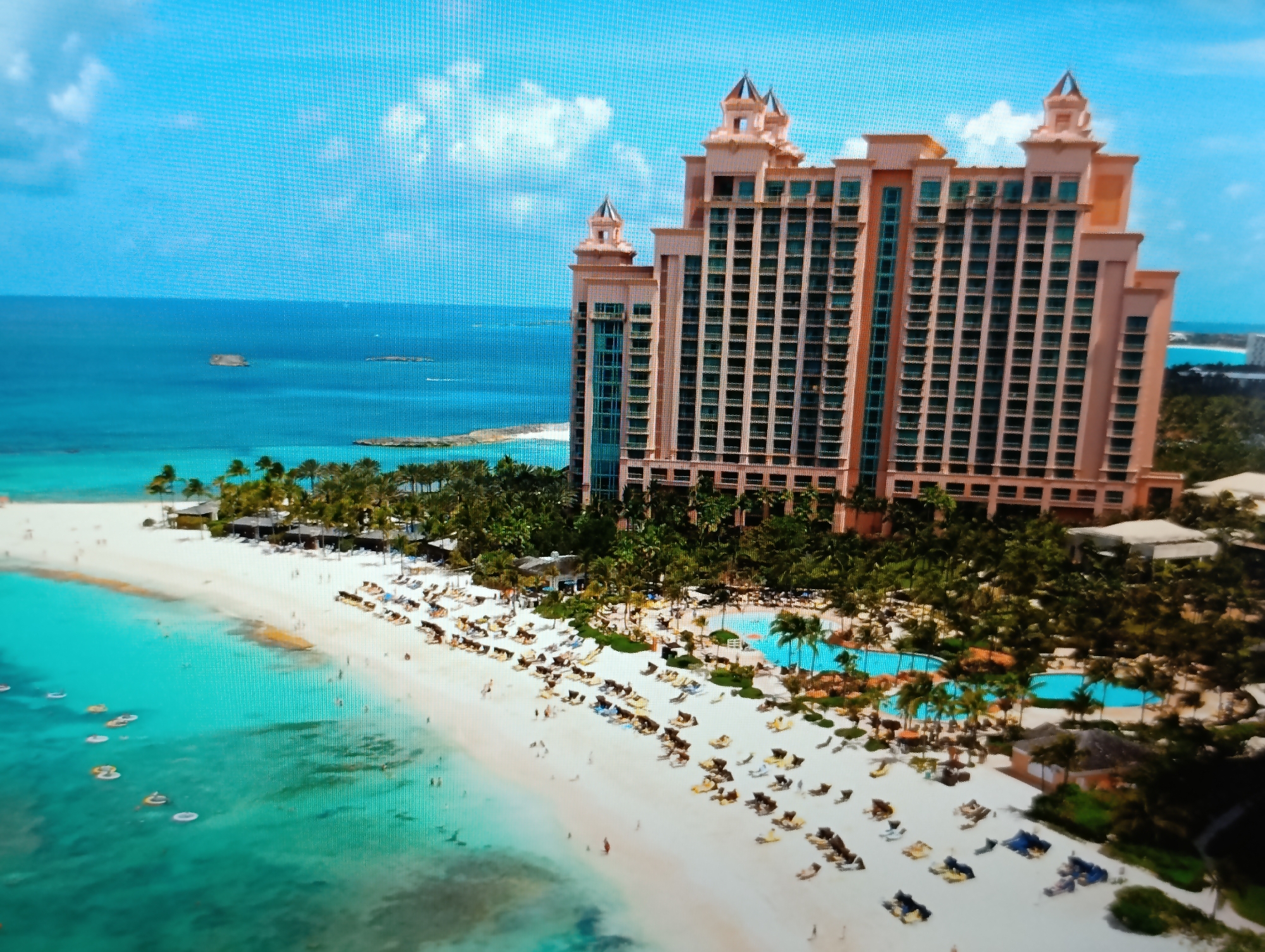 The image depicts a tropical resort scene, with a large, peach-colored hotel building dominating the background. The hotel has a distinctive architectural style with multiple towers and numerous windows. In front of the hotel, a white sandy beach stretches along a clear turquoise ocean. The beach is populated with many lounge chairs and umbrellas, some occupied by people. Palm trees are scattered throughout the resort area, and several swimming pools are visible, filled with blue water. Small inflatable floats are scattered in the shallow waters of the ocean. The sky is blue with scattered white clouds. The scene conveys a sense of a vacation destination. The location is not in Dubăsari town council, Moldova; it is a tropical resort.