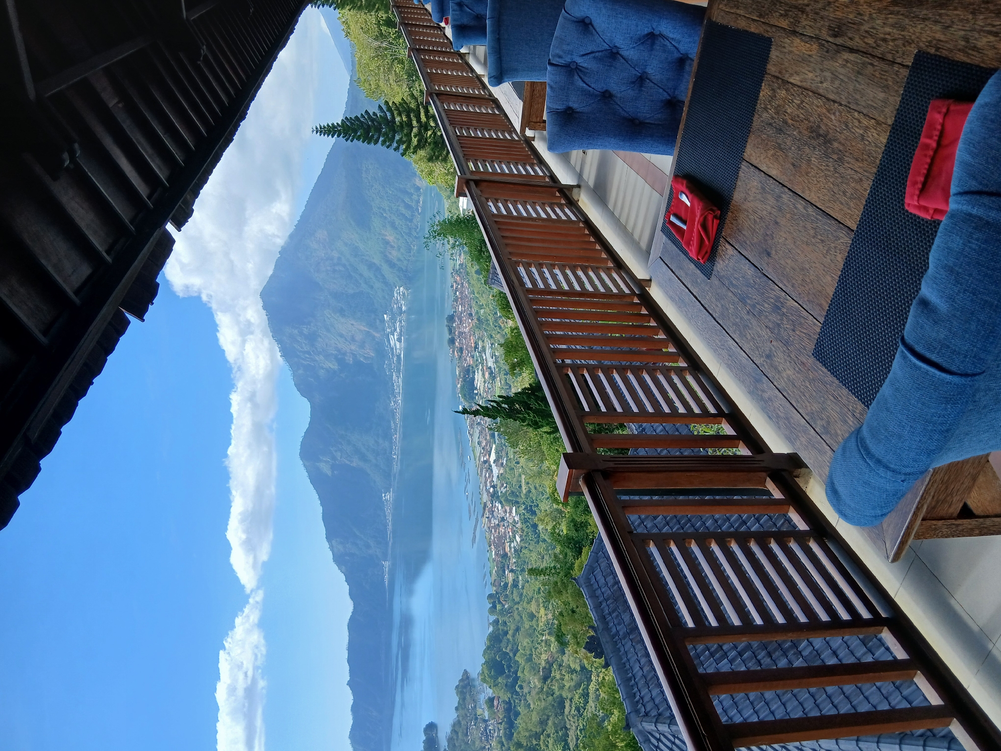 The image shows a scenic view from a covered outdoor dining area, likely a restaurant, overlooking Lake Batur and Mount Batur in Kintamani, Indonesia. The foreground features a rustic wooden table with black placemats and red napkins folded with cutlery. Blue upholstered chairs are visible at the table. A wooden railing with horizontal slats separates the dining area from the view. Beyond the railing, the landscape slopes down to the town surrounding Lake Batur, with a vast expanse of water reflecting the clear blue sky dotted with white clouds. The majestic Mount Batur, a volcano, dominates the background. The architecture suggests a traditional Indonesian style, with a dark wooden ceiling and supporting beams. The overall impression is one of a tranquil and picturesque setting, ideal for enjoying the natural beauty of the region.