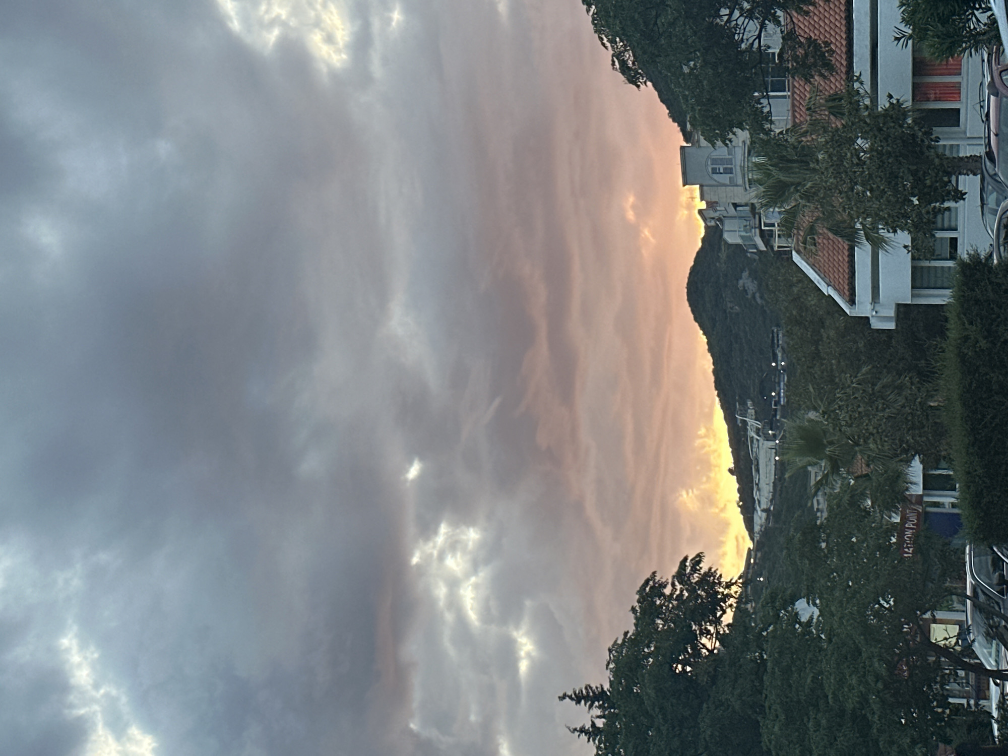 This vertical image depicts a view of Budva, Montenegro, at either sunrise or sunset. The upper two-thirds of the frame are dominated by a cloudy sky, featuring dense, dark grey clouds interspersed with brighter white illuminated sections, particularly in the upper left. Towards the lower horizon, the clouds transition into warm tones of orange, pink, and yellow, indicative of low-angle solar illumination.
In the background, two prominent green, tree-covered mountains or hills rise, framing the illuminated portion of the sky. A golden glow of sunlight is visible behind and between these peaks. On the slope of the mountain on the right, light-colored structures are discernible.
The midground consists of a mix of green foliage and architecture. On the right, a multi-story, light-colored building with multiple balconies and a flat roof is prominent. Adjacent to it, a white building with a reddish-orange tiled roof is partially visible, with a palm tree positioned in front. On the left midground, partially obscured by dense green trees, a red sign with white text reads "MATION POINT".
The foreground features dense green bushes and foliage, which obscure the lower portions of parked vehicles. The visible parts include a dark-colored SUV on the left and a red car on the right. No individuals or explicit human activity are discernible within the frame.