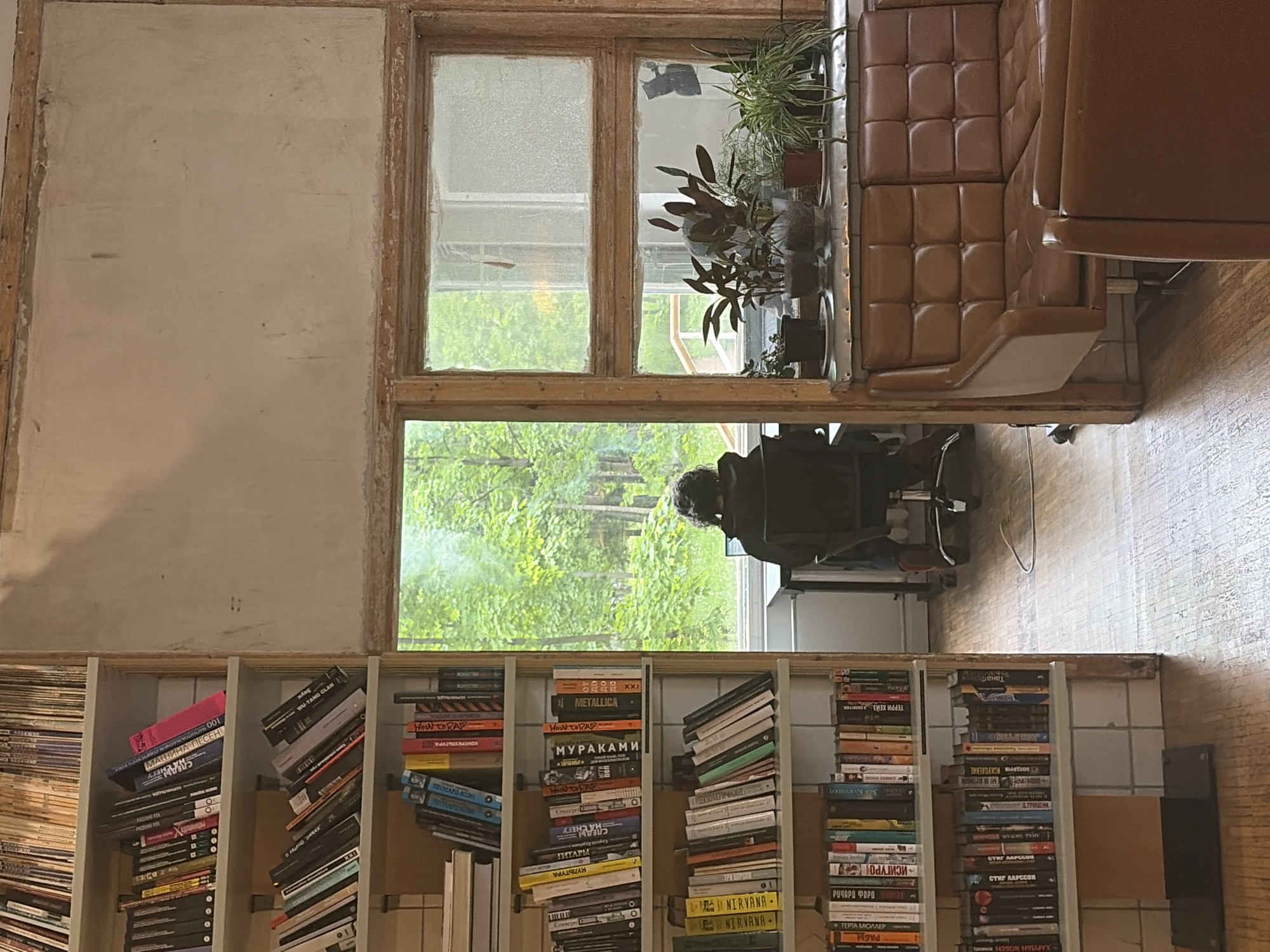 The image shows an indoor scene, likely a study or living room, with an open window providing a view of lush green trees outside. The room is furnished with a brown leather cushioned sofa and a wooden bookshelf filled with books of various colors and sizes. A person is seated near the window, facing the outside. The walls are painted in a neutral color, and the overall atmosphere is calm and serene. The location appears to be a private residence, possibly in a suburban or rural area given the abundance of greenery.