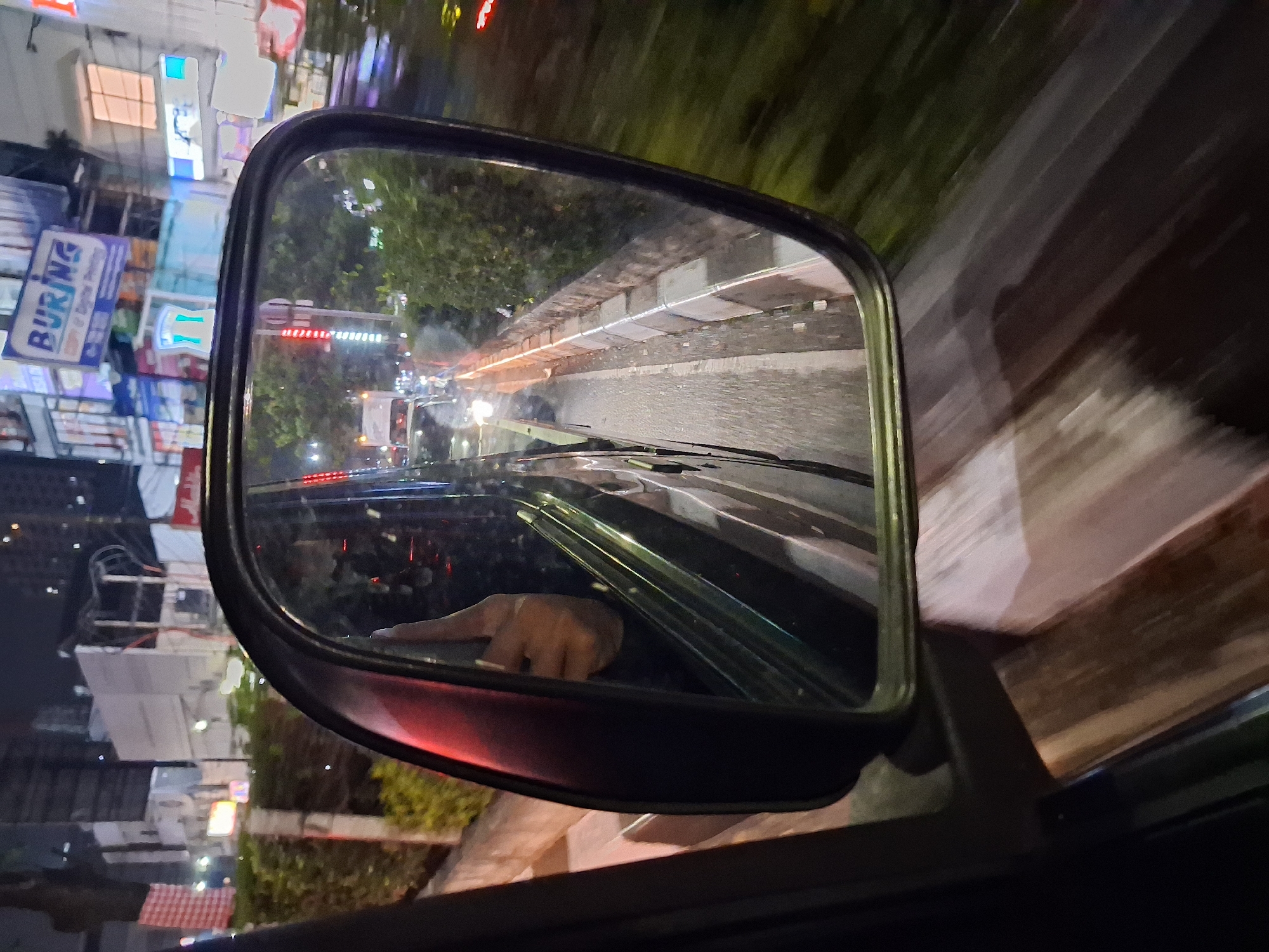The image captures a nighttime view from inside a vehicle in Depok, Indonesia, as seen through the side mirror. The reflection in the mirror shows a hand with a raised thumb, suggesting a gesture of approval or acknowledgement. The surrounding environment reflects a city street scene with blurred lights and foliage. In the background, neon signs and buildings are visible, indicating an urban setting. The street itself appears wet, possibly from rain, with the headlights of other vehicles illuminating the scene. The side mirror is mounted on the exterior of the vehicle, and the angle suggests the photo was taken while the vehicle was in motion. The image is characterized by motion blur, which is typical of photographs taken from a moving vehicle at night.