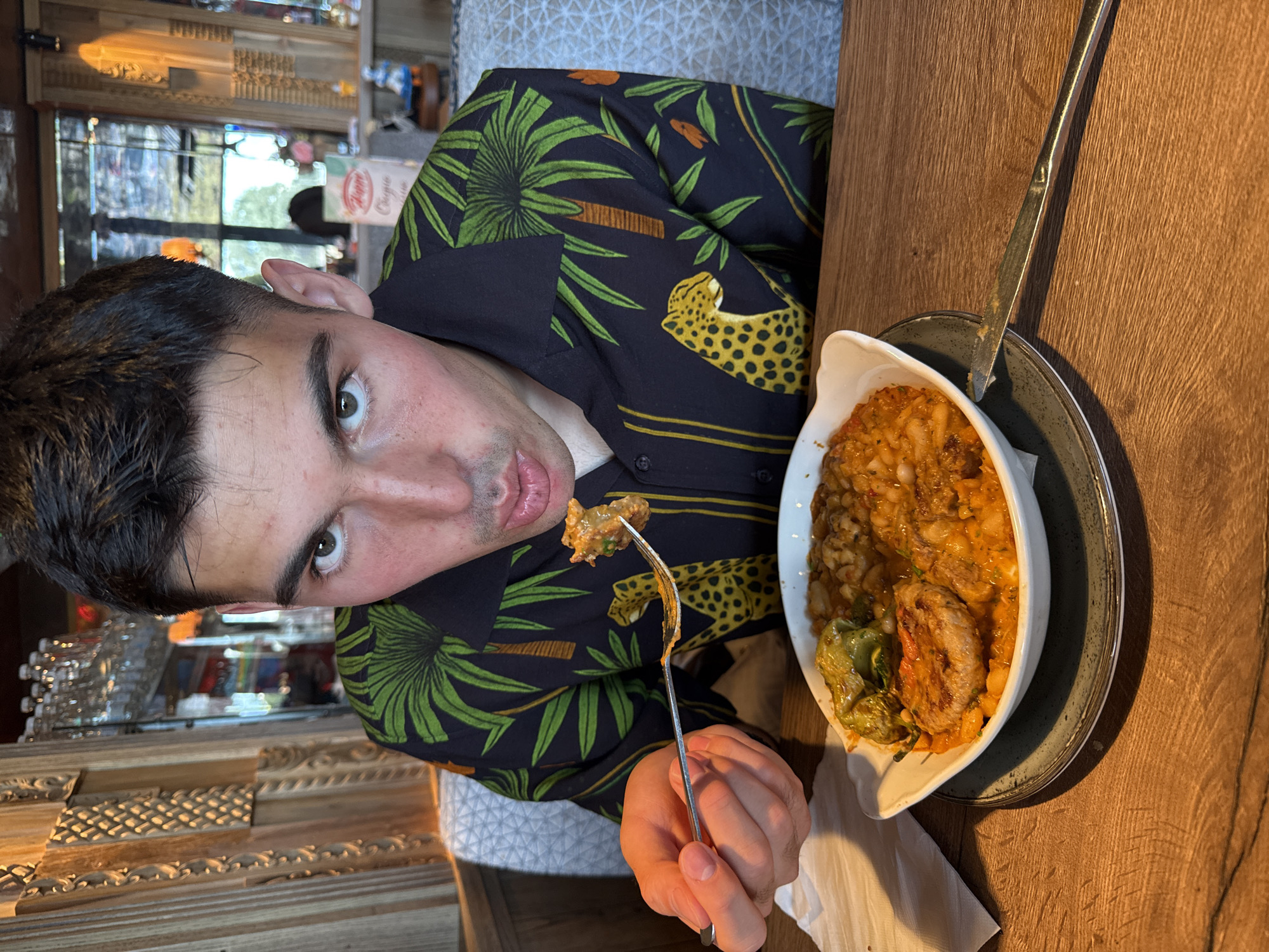 A young man is captured mid-meal in an indoor dining setting, looking directly at the camera with a direct and slightly expressive gaze. He is holding a fork with a bite of food, poised near his open mouth. He wears a dark short-sleeved shirt patterned with vibrant green palm trees, yellow leopards with black spots, and small orange bird-like figures.
In front of him, a white oval dish holds a substantial meal, appearing to be a rich, orange-hued stew made with large white beans, chunks of meat, and green peppers. The dish is placed on a dark-rimmed plate, resting on a warm, wooden table. A metal knife lies beside the plate.
The background suggests a rustic or traditionally styled restaurant interior, with textured wooden paneling and a bar area featuring shelves stocked with various glasses and bottles. Reflections from light sources or windows are visible in the background. A partially obscured red and white sign can be seen, with legible text that appears to be "Cigla" or "Chigla." The overall scene depicts a casual dining experience, set in a restaurant context within Radinovo, Bulgaria.