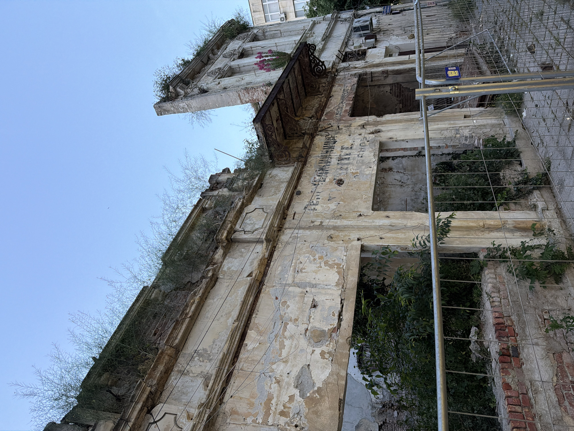 An eye-level upward shot captures the weathered facade of a derelict building in Burgas, Bulgaria. The building is in a state of disrepair, with peeling paint exposing the underlying brickwork and structural elements. Vegetation sprouts from crevices and atop ledges. The windows are vacant and some floors are entirely collapsed, revealing the building's interior overrun with plants. The building's ornate balcony is corroded. Adjacent to the building, a temporary wire mesh fence stands, supported by metal poles. A small blue sign is attached to the fence. The sky is a clear, light blue.