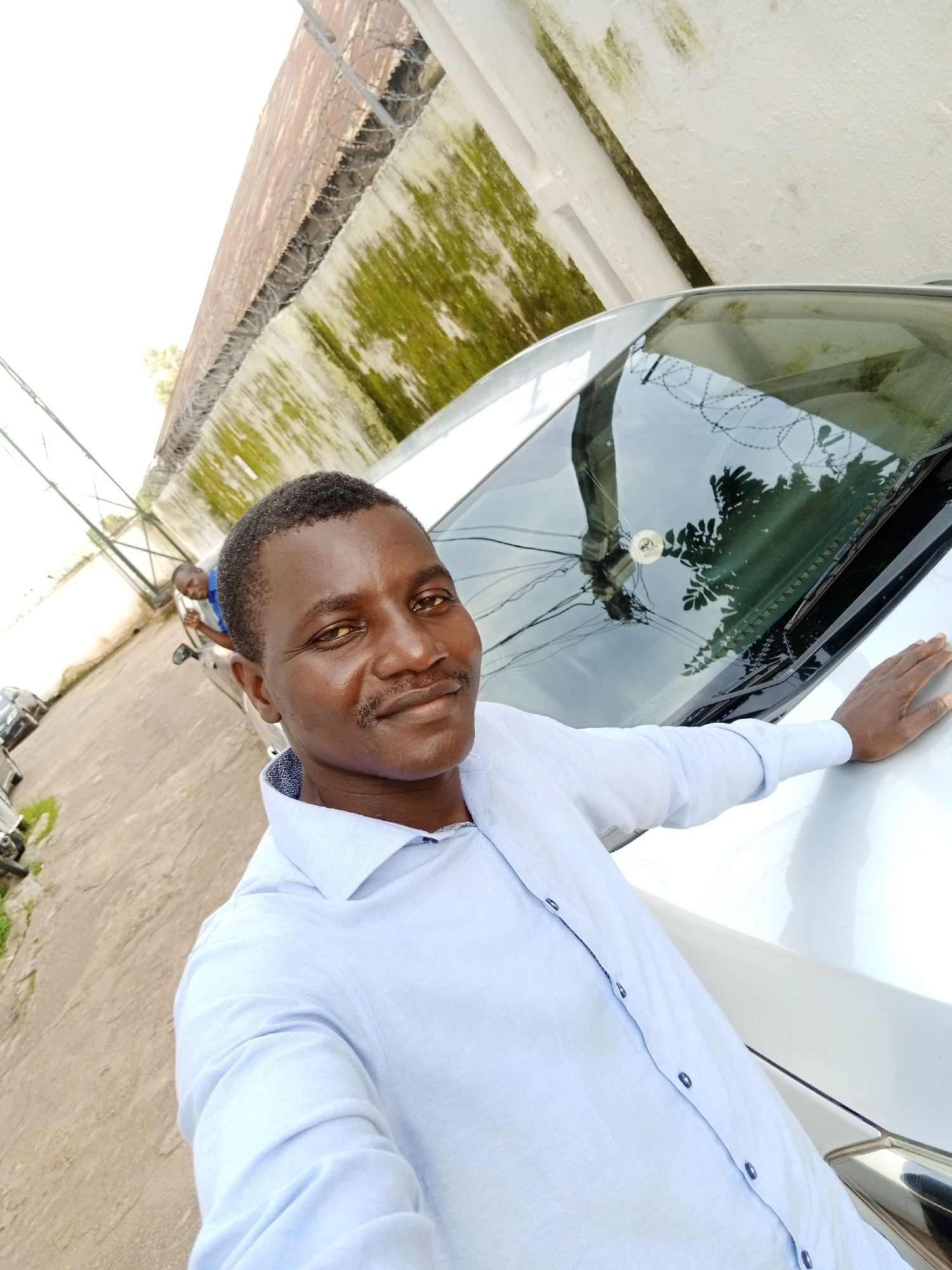 A man in a light blue collared shirt is taking a selfie next to a white car. His right hand is resting on the hood of the car. In the background, another man in a blue shirt is leaning out of a car door. The scene appears to be in an outdoor parking lot with a weathered concrete ground. Behind the cars, a textured wall with green moss and a corrugated metal fence topped with barbed wire is visible. The overall lighting suggests it is daytime. The location is identified as Jos, Nigeria.