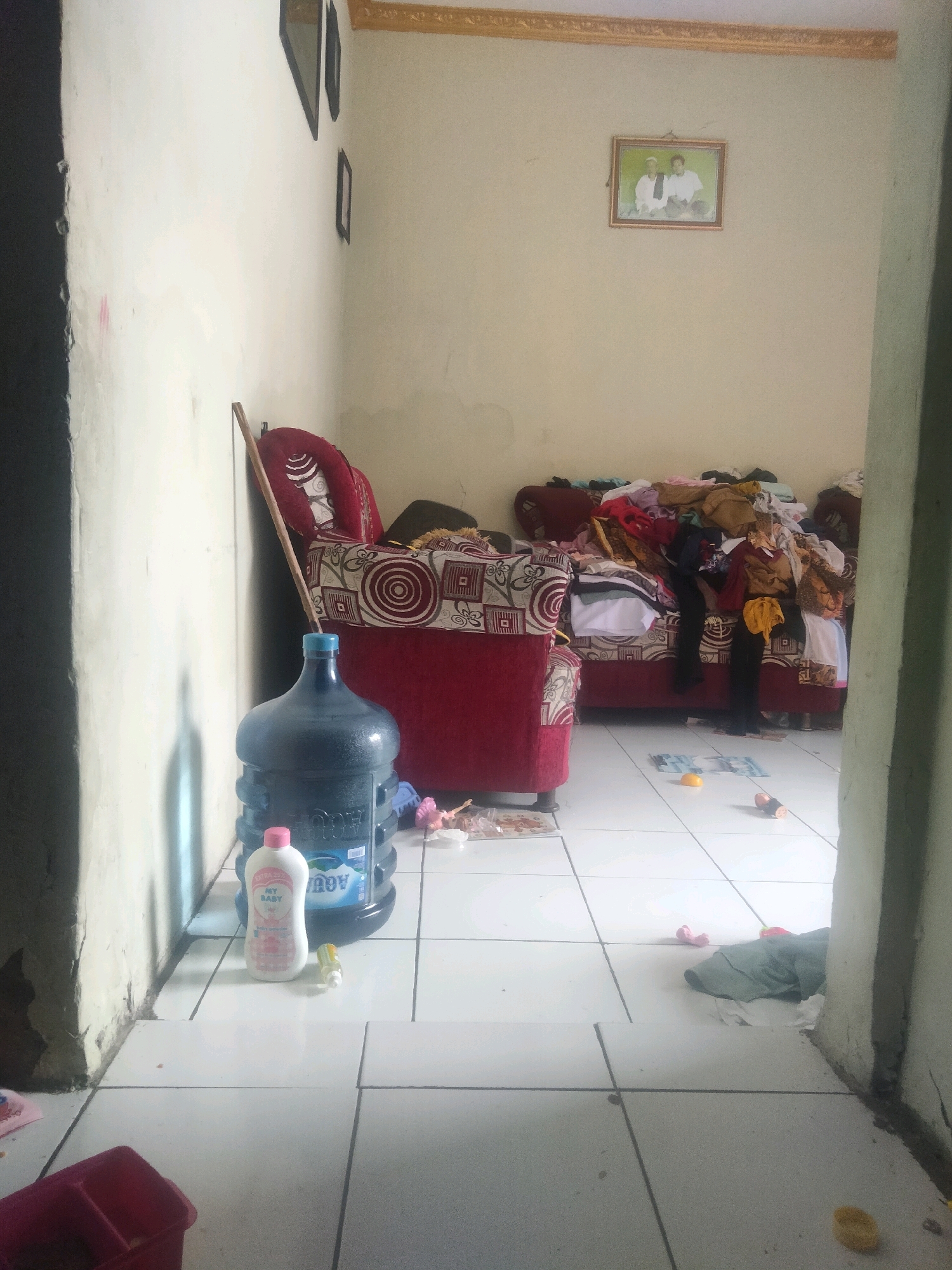 The image depicts a cluttered interior scene, likely a living room or common area, in Suka Sari, Indonesia. In the foreground, a large blue water dispenser jug stands on a white tiled floor, accompanied by a white bottle of "MY BABY" baby powder. To the left, a portion of a white wall with some discoloration and a black-painted edge is visible. A wooden stick leans against this wall. Further into the room, a red armchair with a patterned fabric is positioned. Beyond the armchair, a couch is heavily laden with a large pile of assorted clothing and textiles, creating a disorganized appearance. Several framed pictures hang on the beige wall in the background, including one at the top right corner featuring two individuals. The floor is a grid of white tiles, with various small objects scattered across it, including what appears to be a yellow toy, some currency, and other unidentifiable items. The overall impression is one of a lived-in space that is untidy.