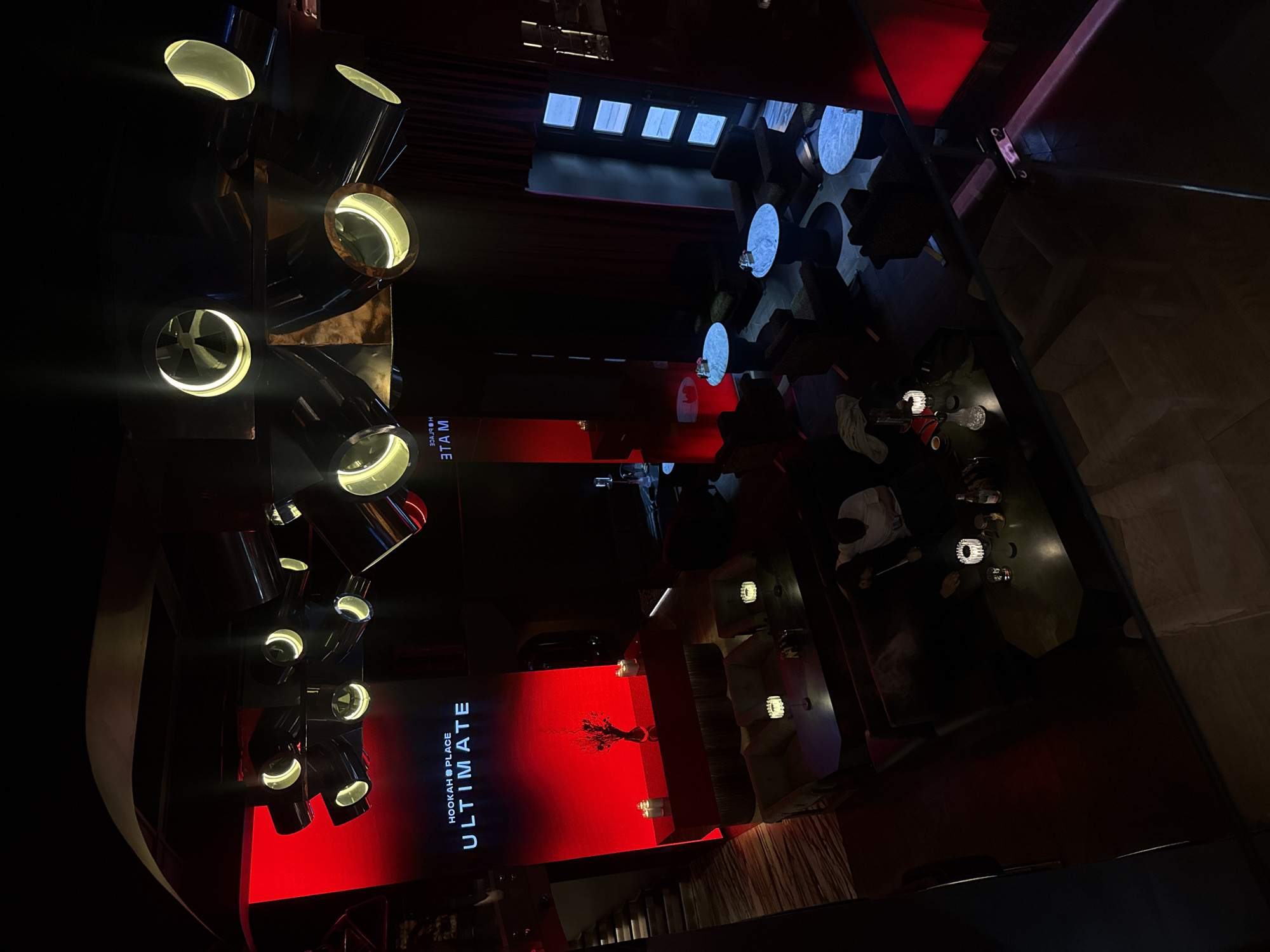 This image captures the interior of a modern, dimly lit lounge or club, likely during the evening or night, in Moscow, Russia. The perspective is from a slight elevation, looking down into the space.
On the left, a prominent multi-component lighting fixture is visible, featuring several large, cylindrical metallic spotlights with bright, glowing white-yellow interiors, casting beams of light into the dark room. Below these lights, a wall is dramatically illuminated with a vibrant red light, serving as a backdrop for a screen. This screen displays clear white text: "HOOKAH PLACE" and "ULTIMATE," along with a stylized logo, possibly depicting a stag or deer head.
In the central and right areas of the image, several patrons are seated at round tables with reflective surfaces, surrounded by dark, plush chairs or banquettes. They appear to be socializing. Glasses and bottles are visible on some tables, and a hookah is discernible on one. The overall ambiance is sophisticated and atmospheric, created by the low lighting and strategic use of bright, contrasting illumination. Dark architectural elements, such as pillars and ceiling structures, are present, along with what appear to be curtained sections or dark drapes in the upper background. Reflections, including the reversed word "MATE" (ЭТАМ), are visible on shiny surfaces. The setting suggests a venue designed for social gatherings and entertainment.