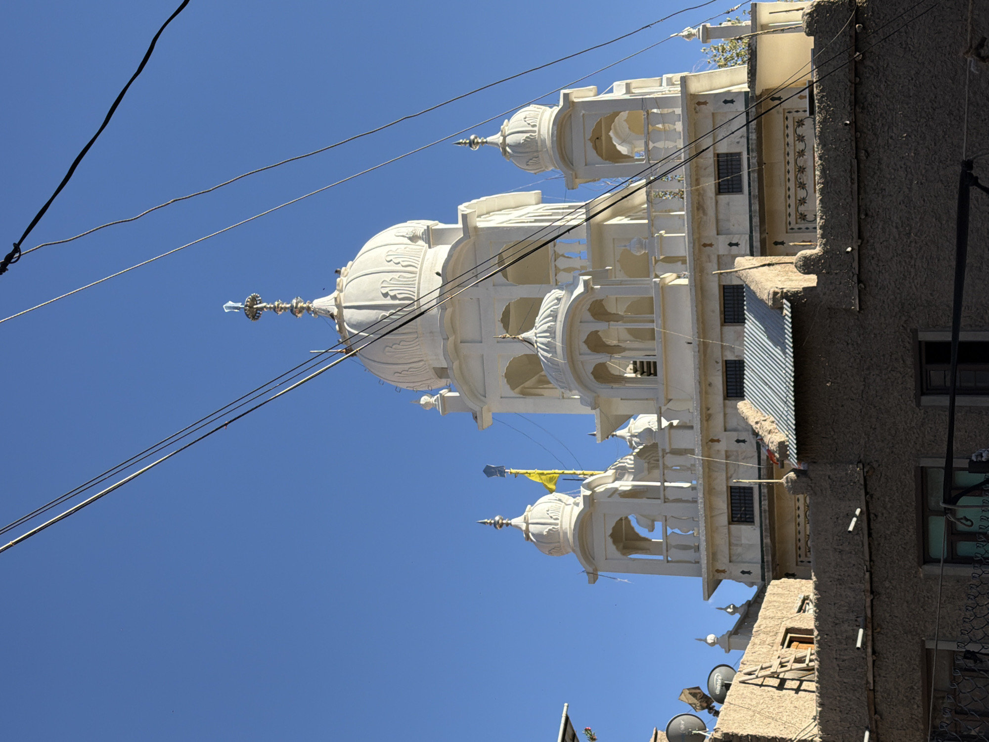 The image shows a white Sikh Gurdwara, likely located in Leh, India, with a clear blue sky as the background. The Gurdwara features a prominent dome, intricate architectural details, and several smaller domes and minarets. In the foreground, there are lower, mud-colored buildings and a few satellite dishes on one of them. Several electrical wires run across the frame, obscuring parts of the sky and the Gurdwara. The overall scene suggests a sunny day in an urban or semi-urban environment with a mix of traditional and modern elements.